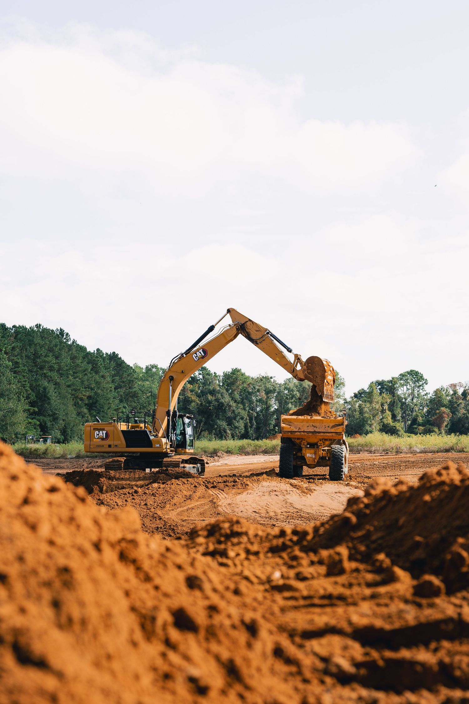 Two construction excavators moving dirt at an active construction site with trees in background.