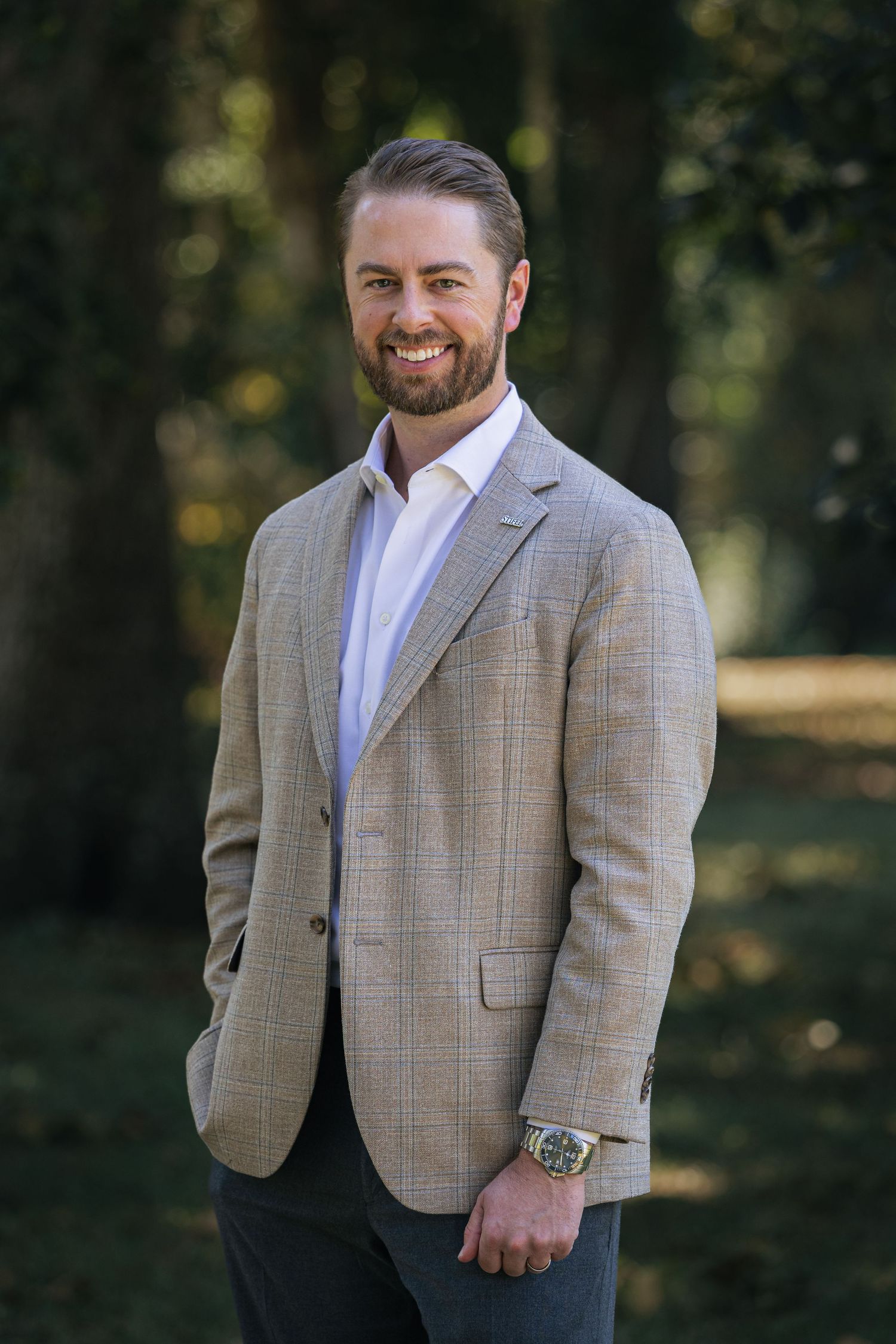 A person wearing a plaid grey suit jacket and white dress shirt poses outdoors against a blurred forest background.