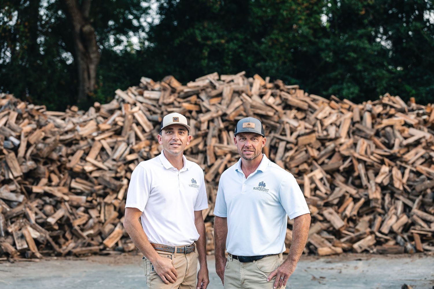 A large pile of chopped and stacked firewood fills the background while two people stand in casual attire outdoors.