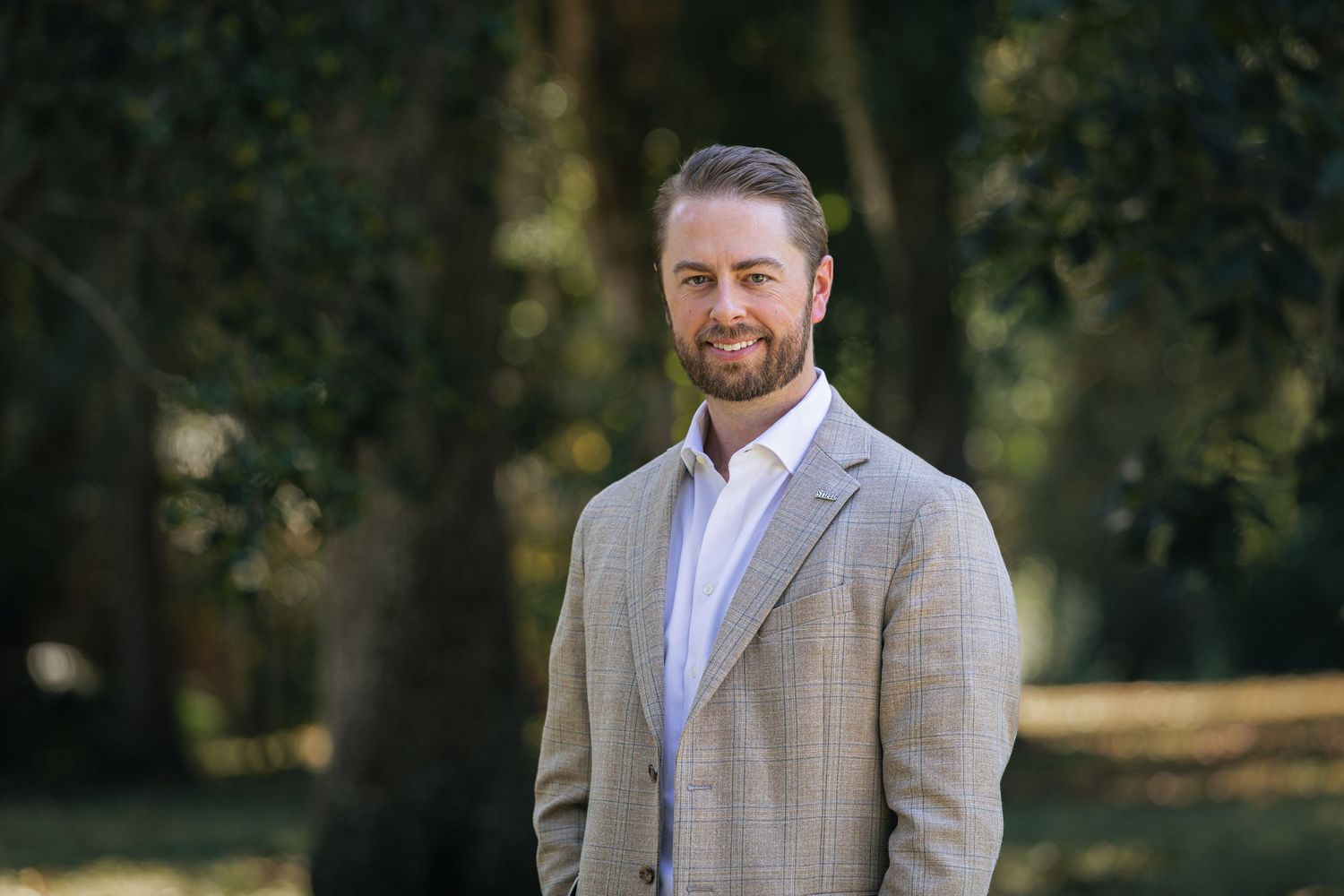 Professional headshot of a businessman in a light gray suit jacket against a blurred outdoor background.