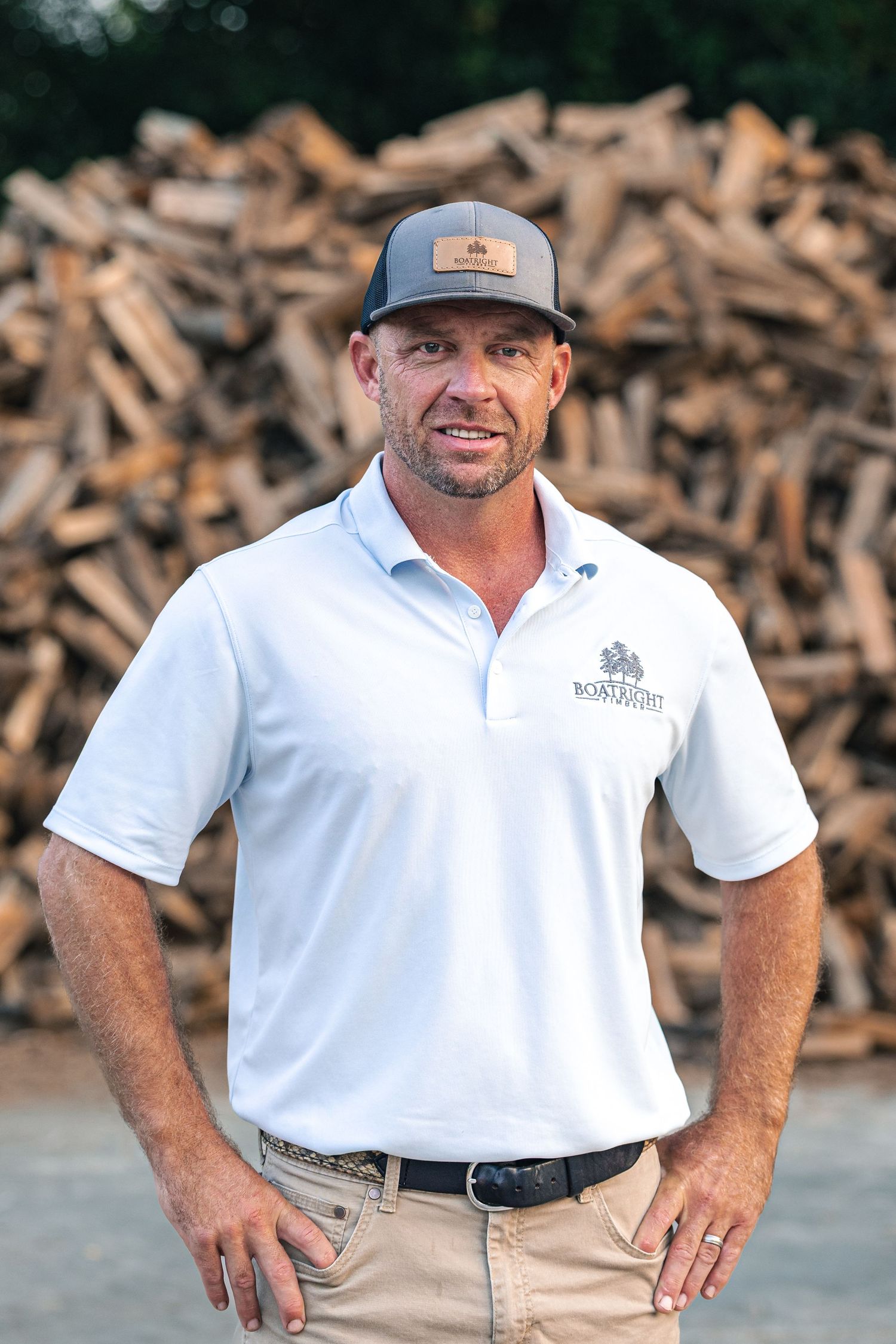 Person wearing white polo shirt and baseball cap smiling outdoors near woodpile.