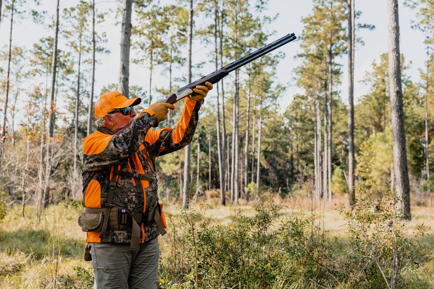 Hunter in orange safety vest aiming rifle while standing among tall pine trees during hunting season.
