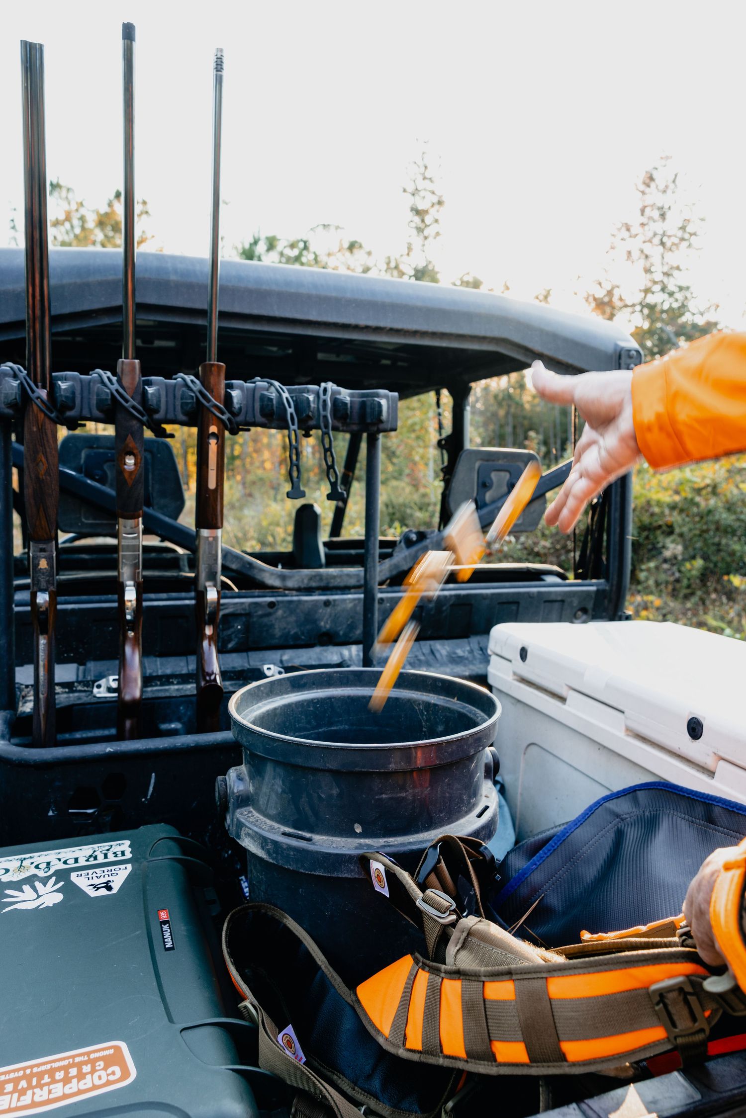 Person in orange jacket loading tools into a bucket on an off-road UTV with rifle rack in outdoor setting.
