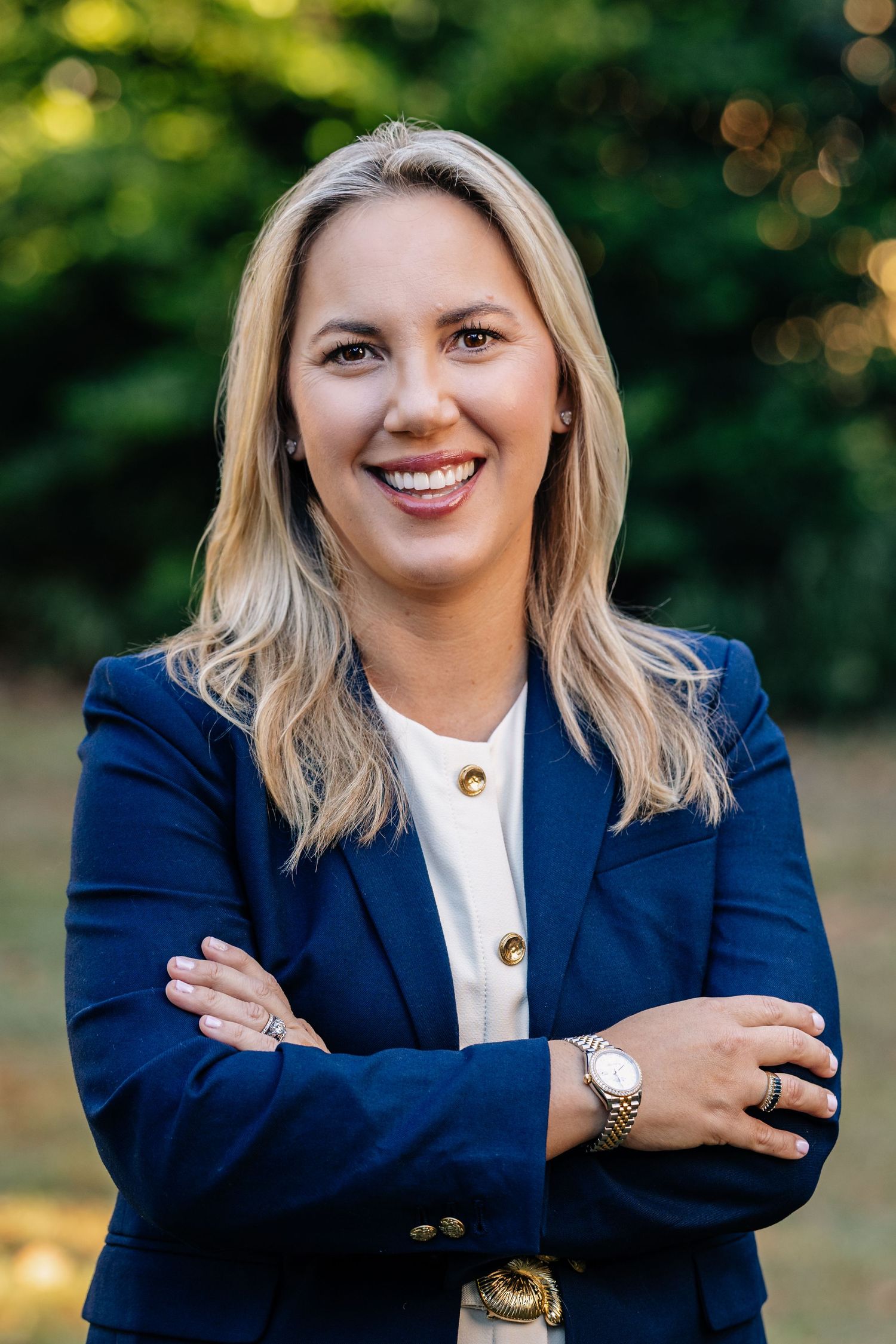 Professional portrait of a businesswoman in a navy blazer and white blouse standing outdoors with arms crossed.