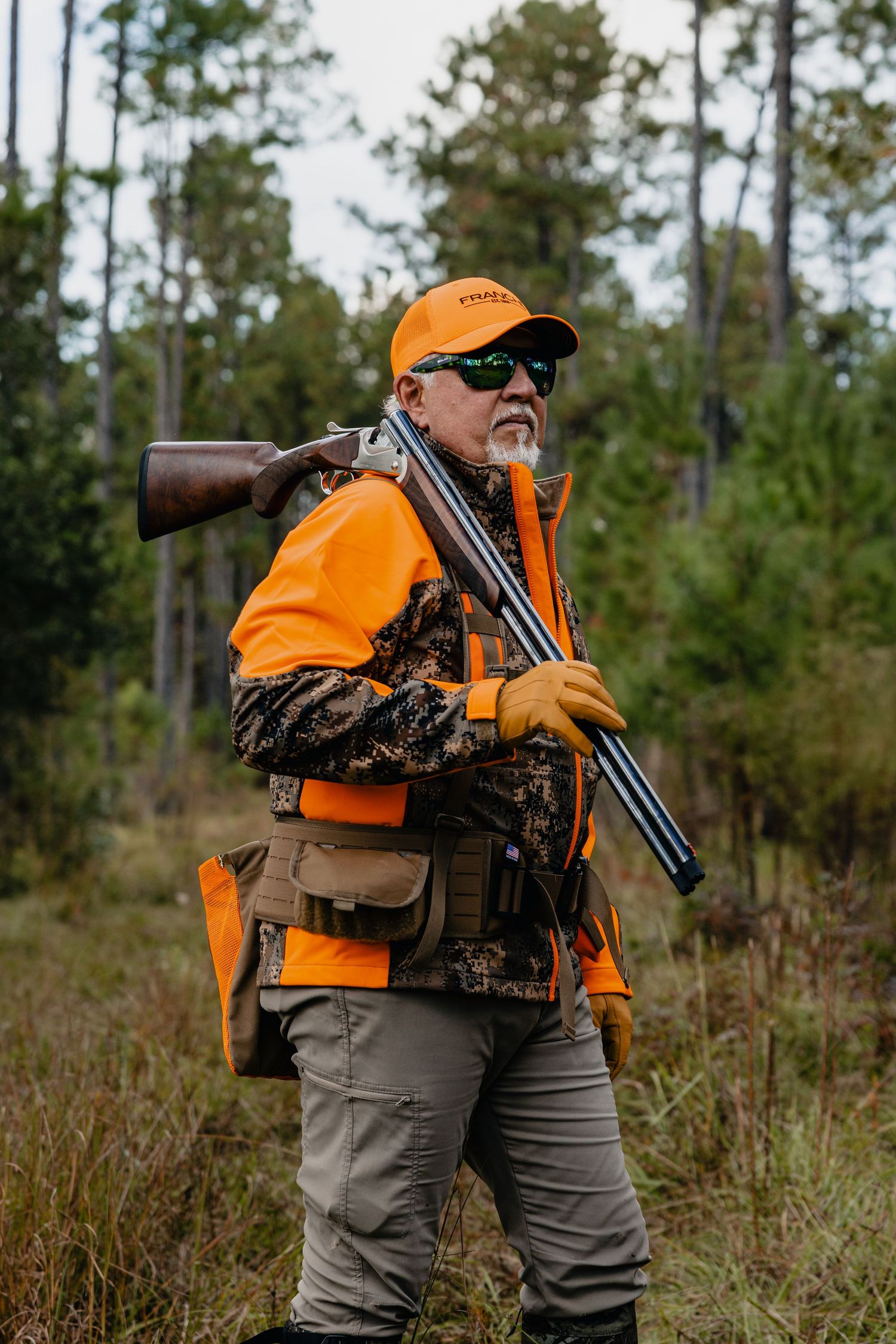 Hunter in orange safety vest and cap carrying a rifle in a wooded forest area during hunting season.