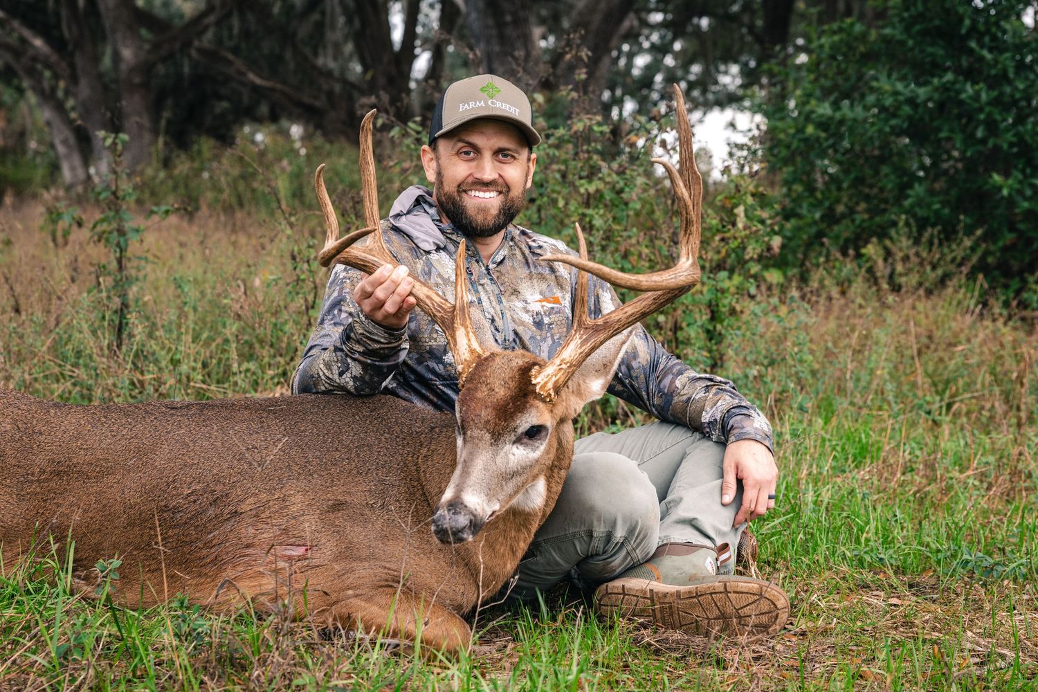 A hunter in camouflage gear poses with a harvested whitetail deer in a grassy field.
