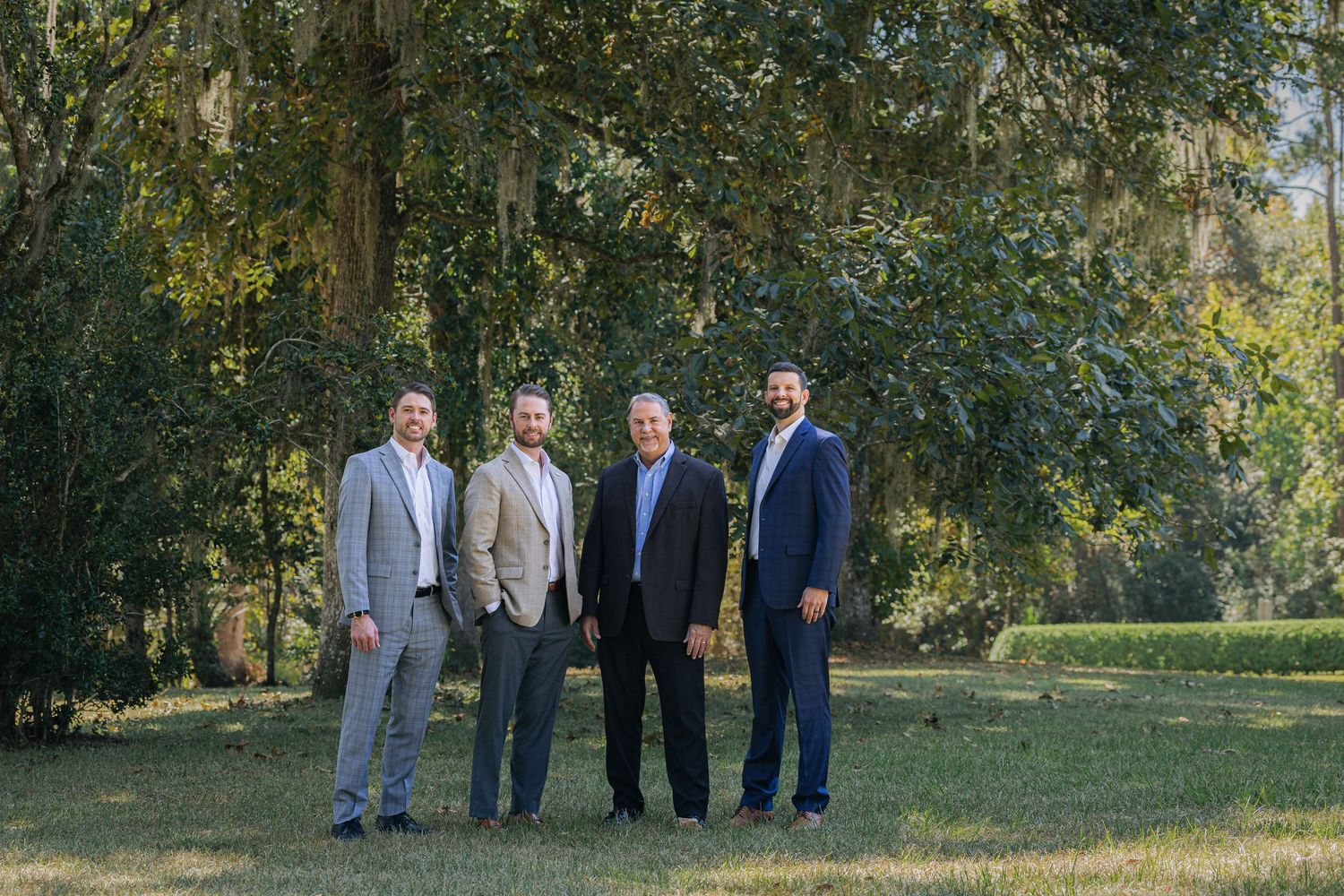 Group of four businessmen in suits standing together outdoors in a wooded setting.