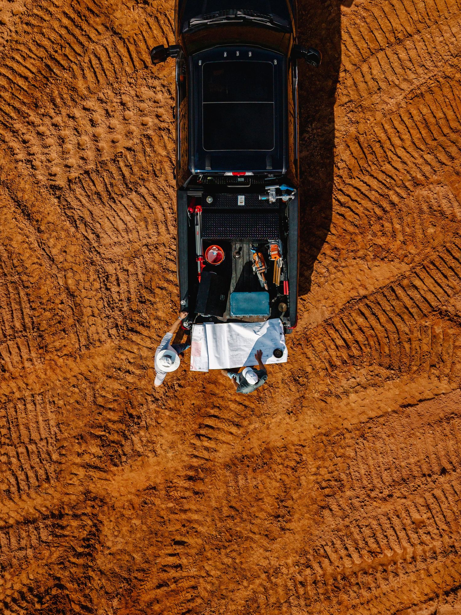 Overhead view of black vehicles on reddish-brown dirt terrain creating tire tracks.