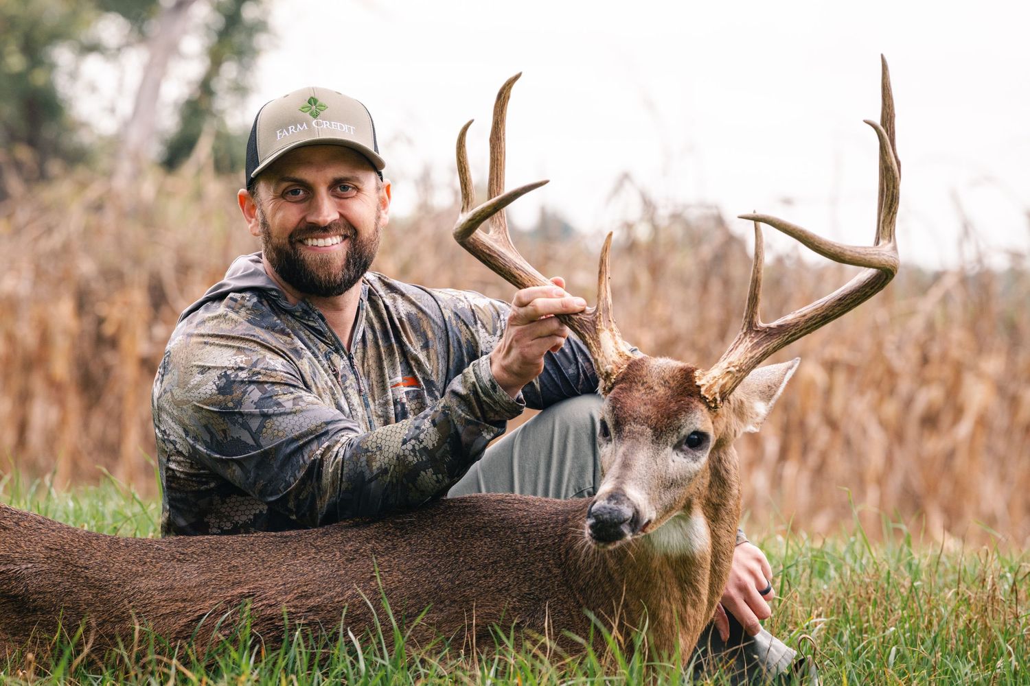 A hunter in camouflage gear poses with a harvested whitetail deer in a grassy field.