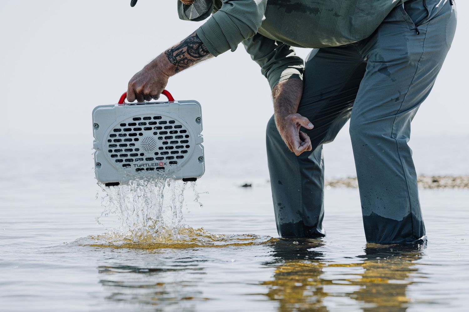 Person in waders releasing bait from metal container into shallow water.