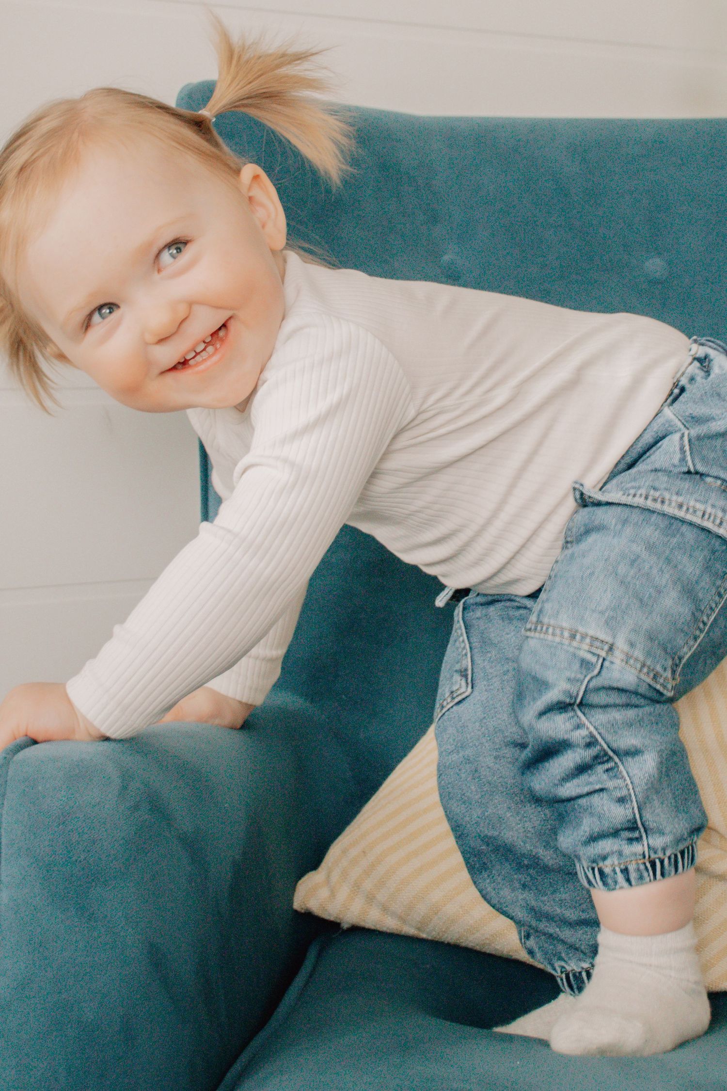 Series of five photos showing a small child in white sweater and jeans playing and moving on a teal couch.