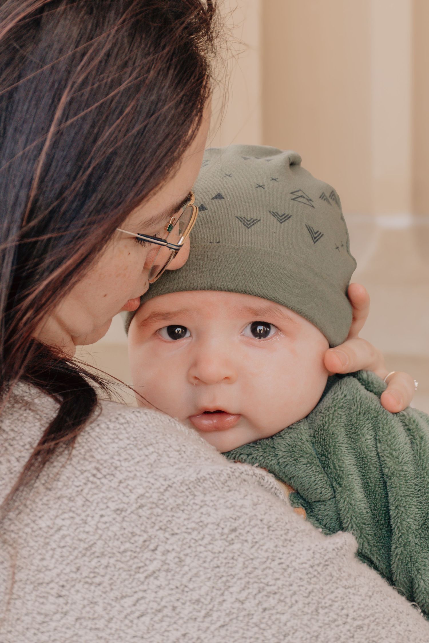 A baby wearing a green beanie and sweater snuggles close to someone in a cozy gray sweater.