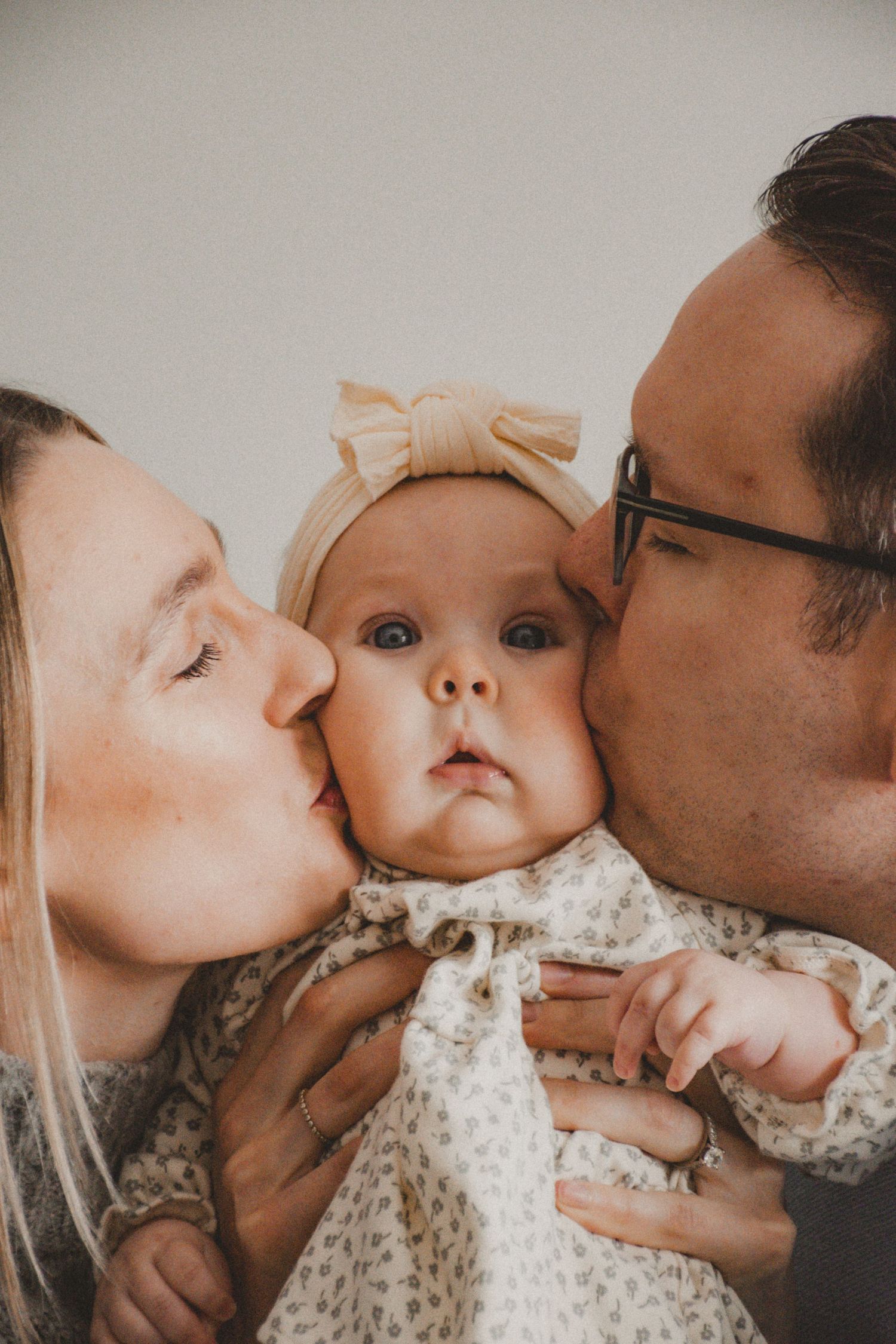 Parents sharing tender kisses with baby wearing beige bow headband and floral dress while taking family photo.
