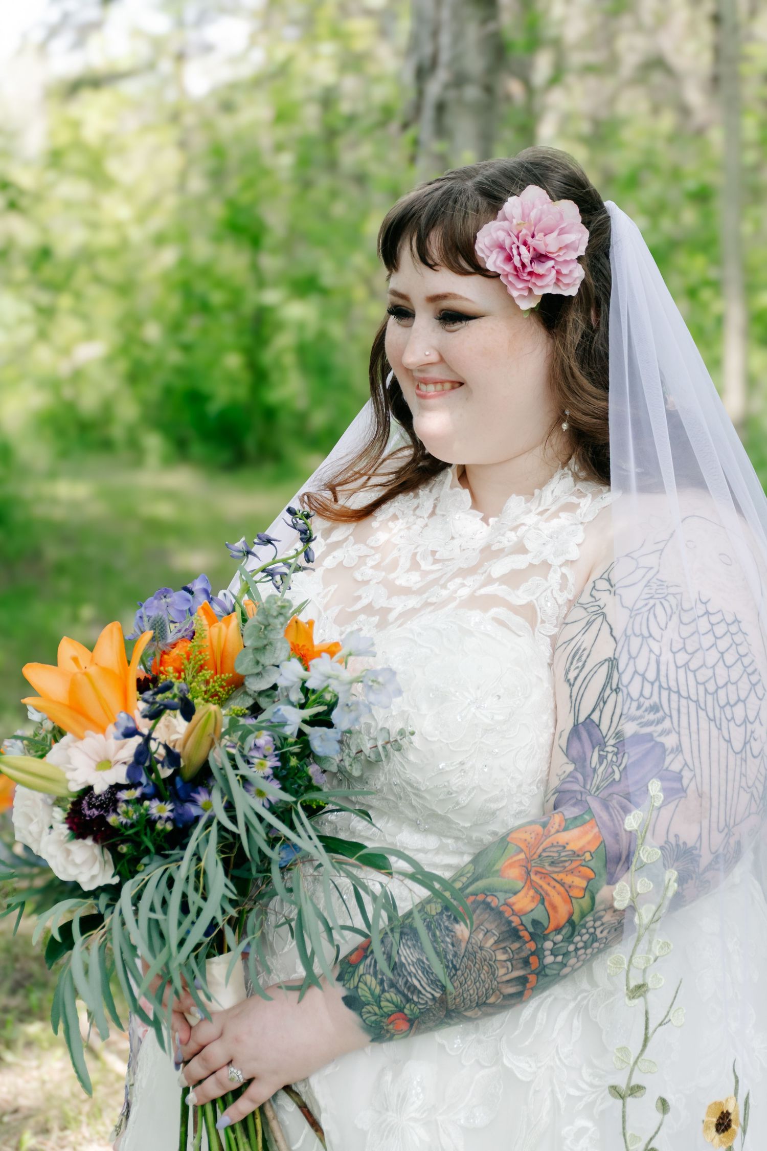 A bride in a white lace dress with colorful tattoo sleeves holds a vibrant bouquet of lilies and wildflowers.