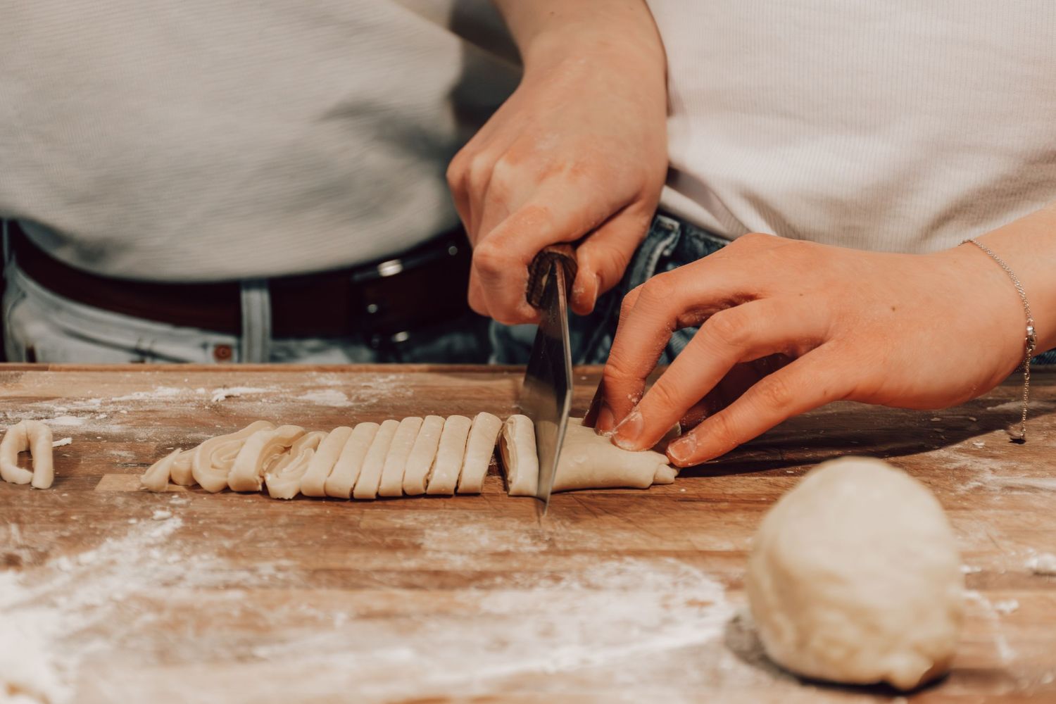 Close-up of hands cutting strips of raw dough on a floured wooden surface with a chef's knife.