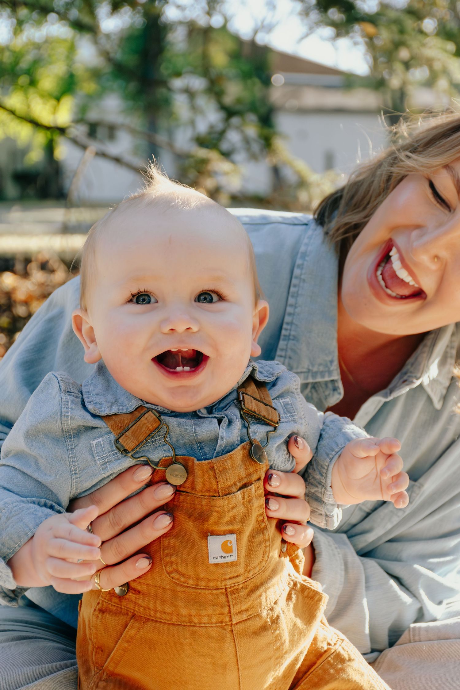 Baby in brown overalls and denim shirt laughs joyfully while being held during outdoor photoshoot.