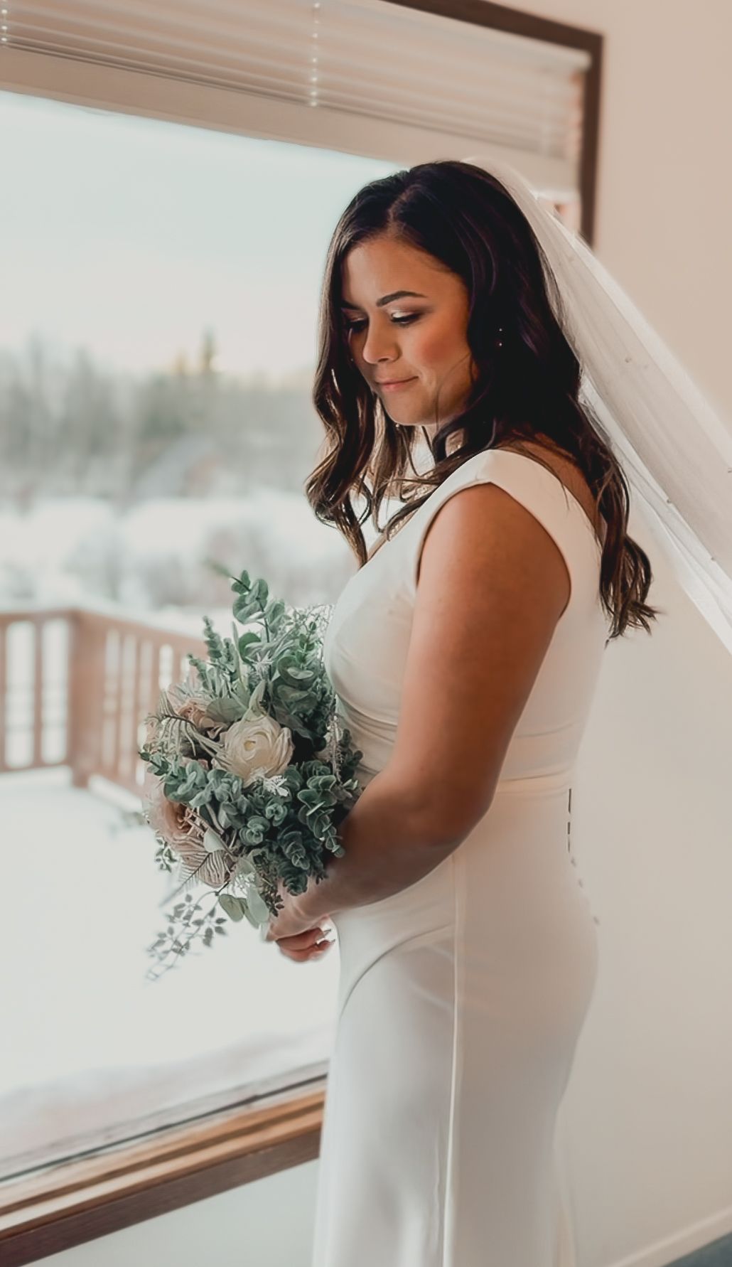 A bride in an elegant white fitted dress holds a delicate bouquet of eucalyptus and white roses by a window.