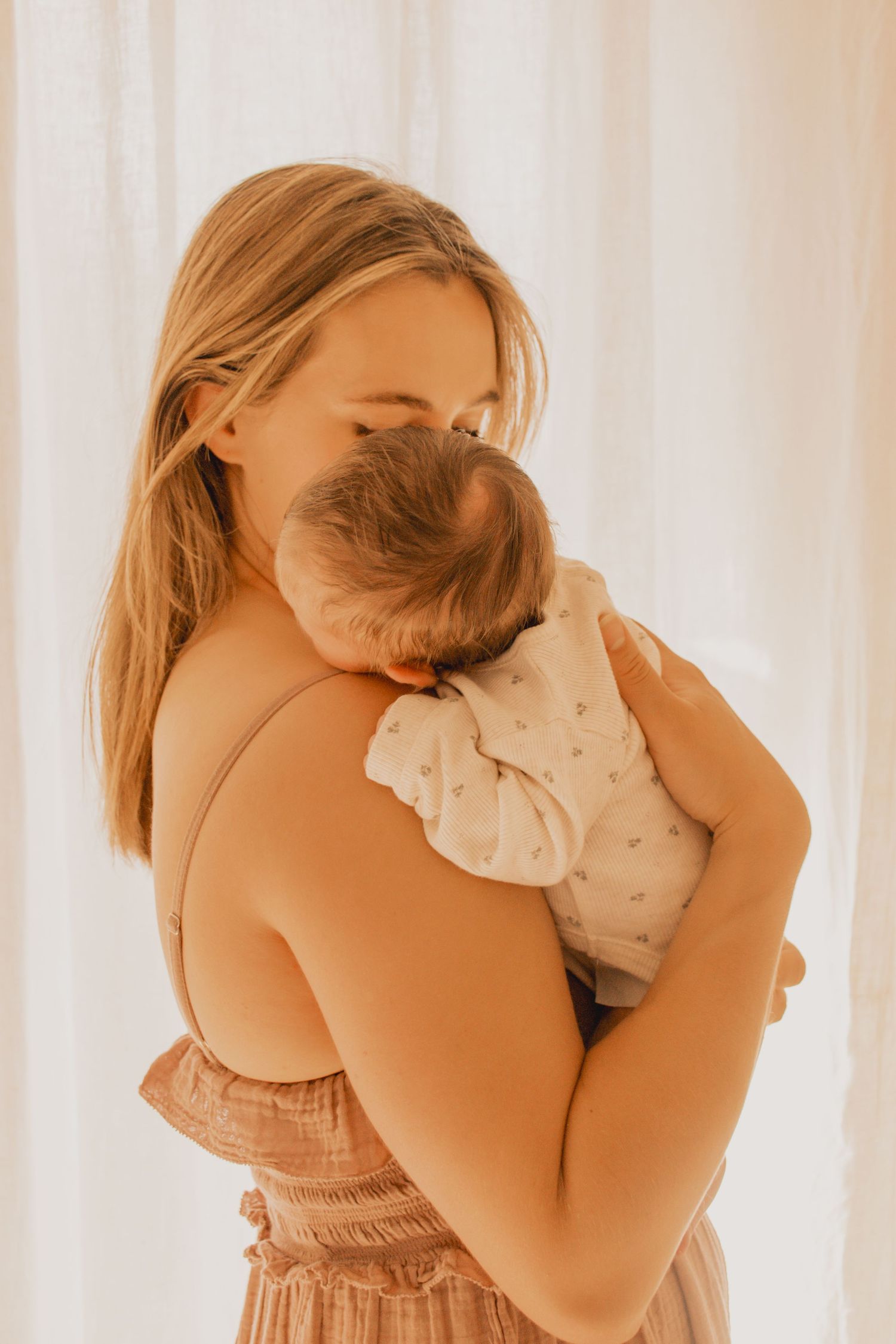 A tender moment captured as mother embraces baby in a sunlit room with white curtains and soft warm tones.
