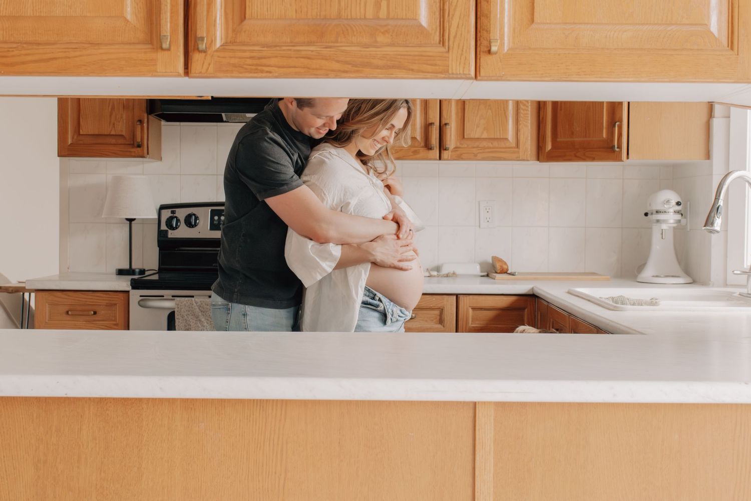 A couple embraces while standing in a kitchen with wooden cabinets and stainless steel appliances.