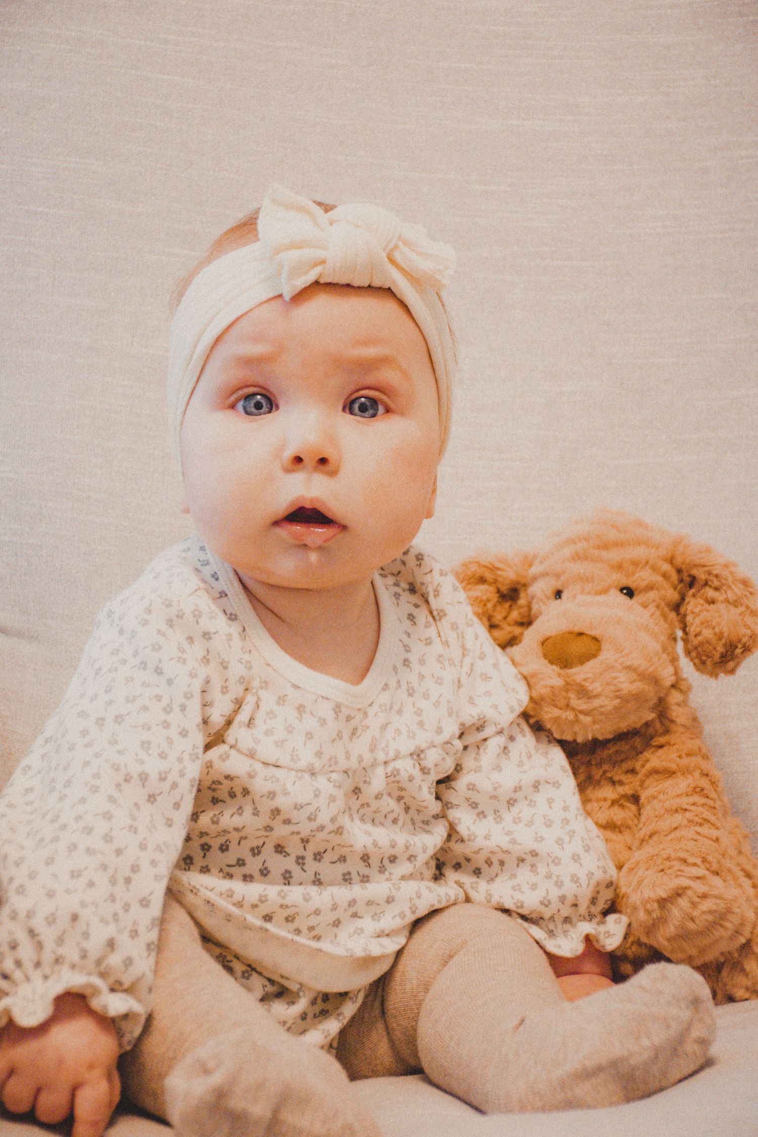 A baby wearing a cream floral dress and bow headband sits with a plush brown teddy bear against a neutral background.