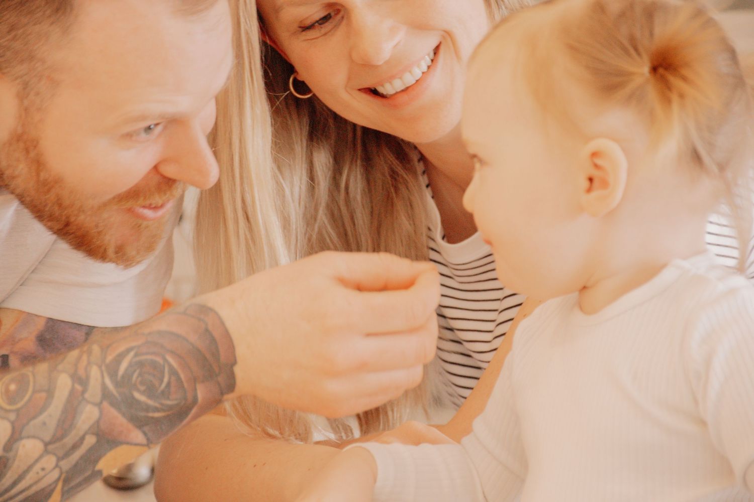A tattooed person shares a warm family moment indoors in golden lighting with a child wearing a striped shirt.