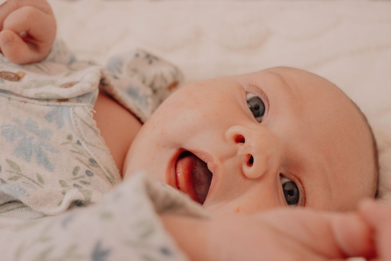 A baby lies on white bedding with floral patterns while making various facial expressions in a series of photos.