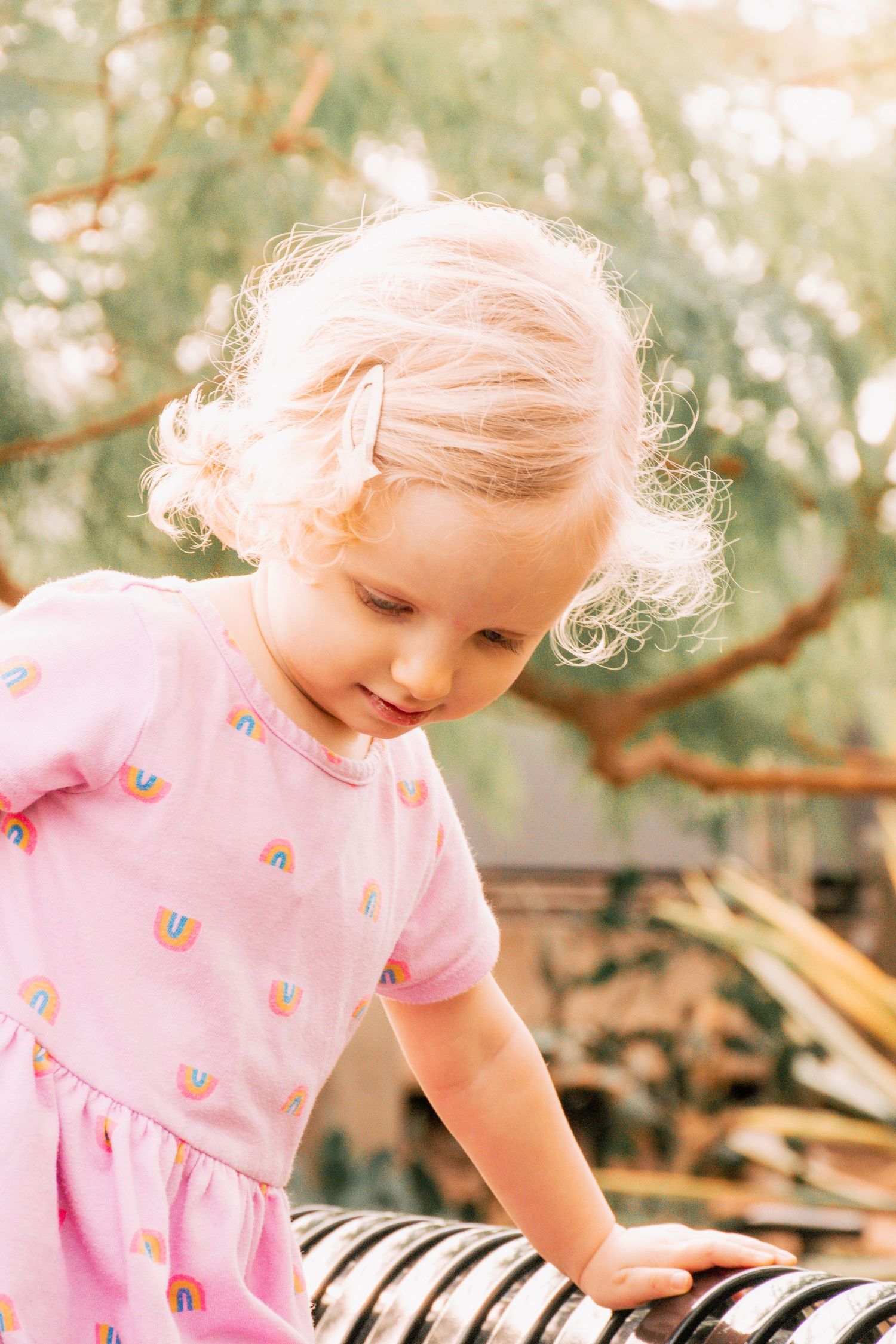Small child wearing pink rainbow dress looking down while standing outdoors in natural lighting.