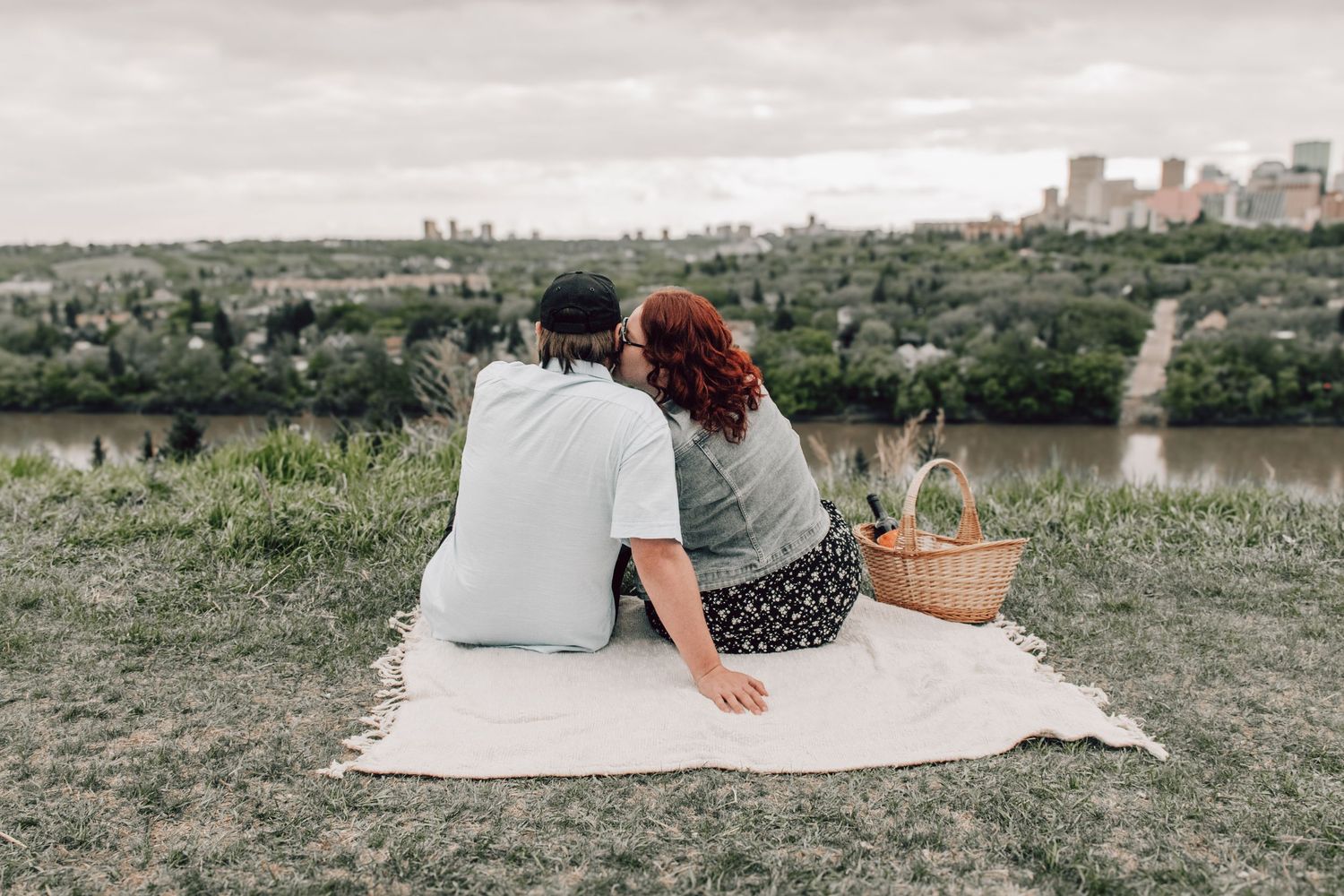 A couple enjoys a picnic on a blanket overlooking a scenic city skyline and green parkland.