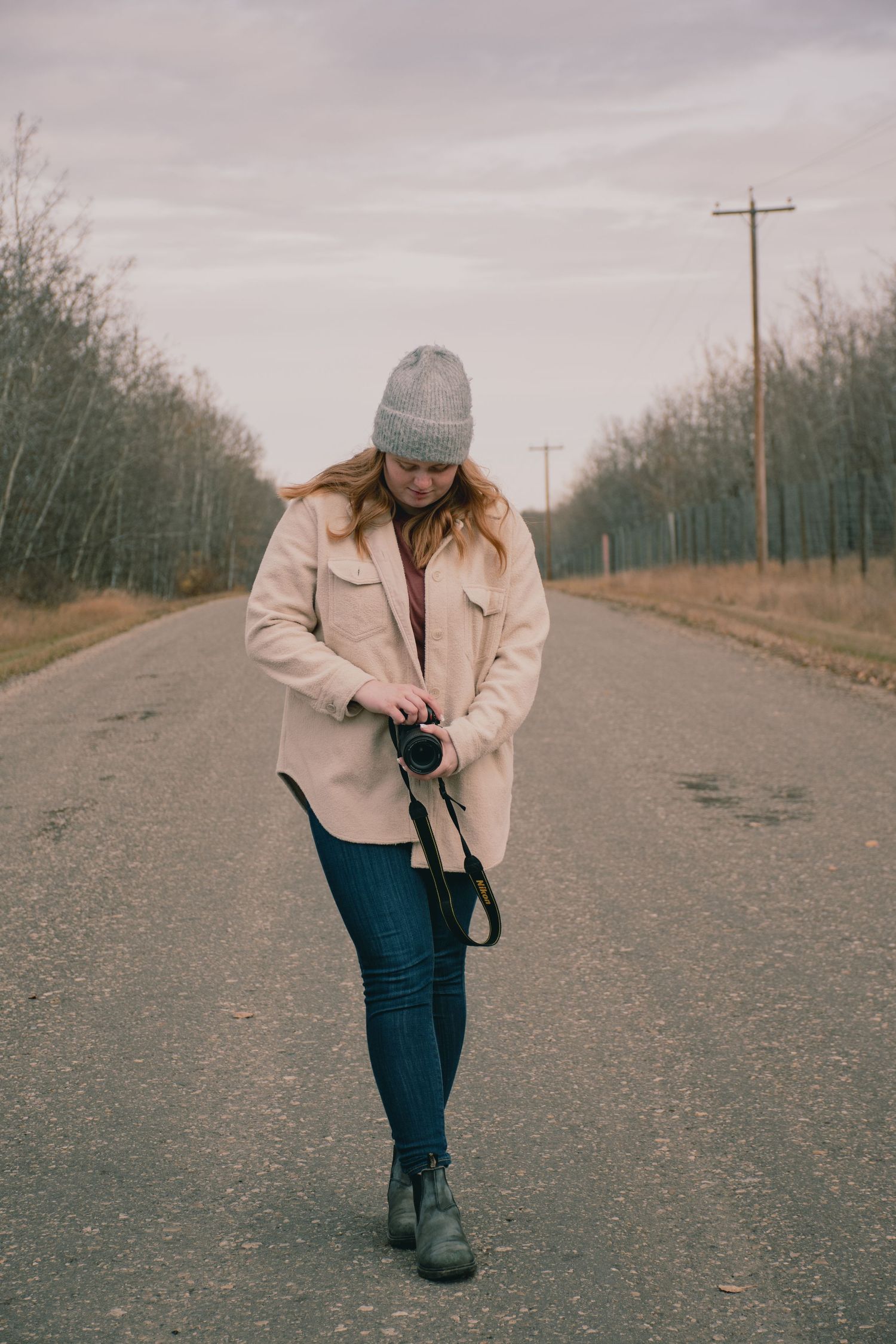 A person in a beige jacket walks down a rural road while looking at their camera.
