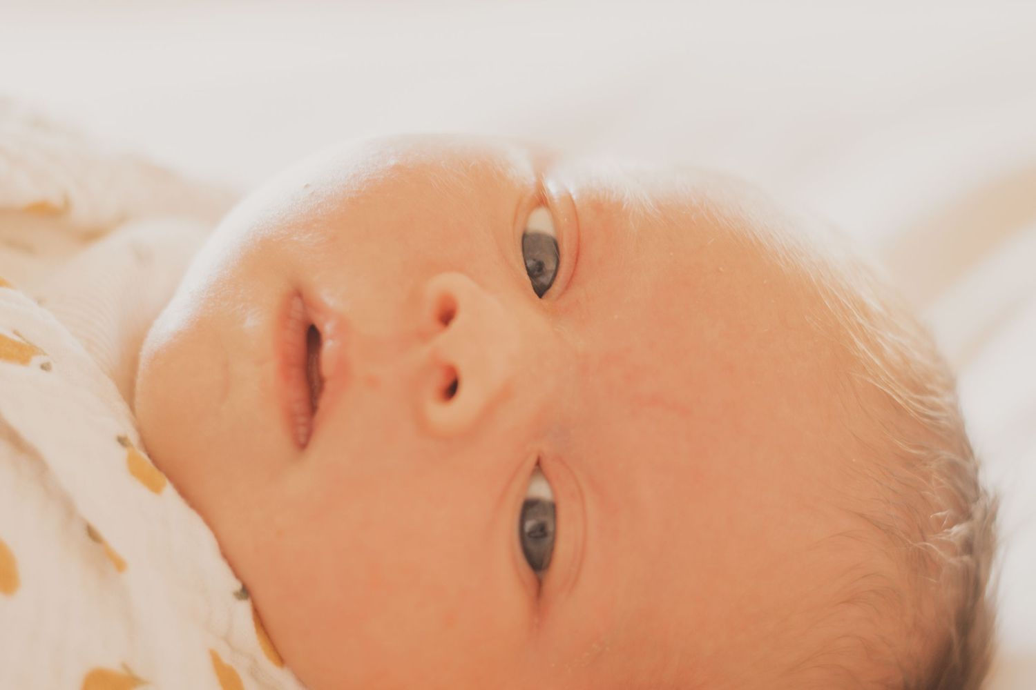 A small baby laying down with a sweet expression wearing a light colored onesie.