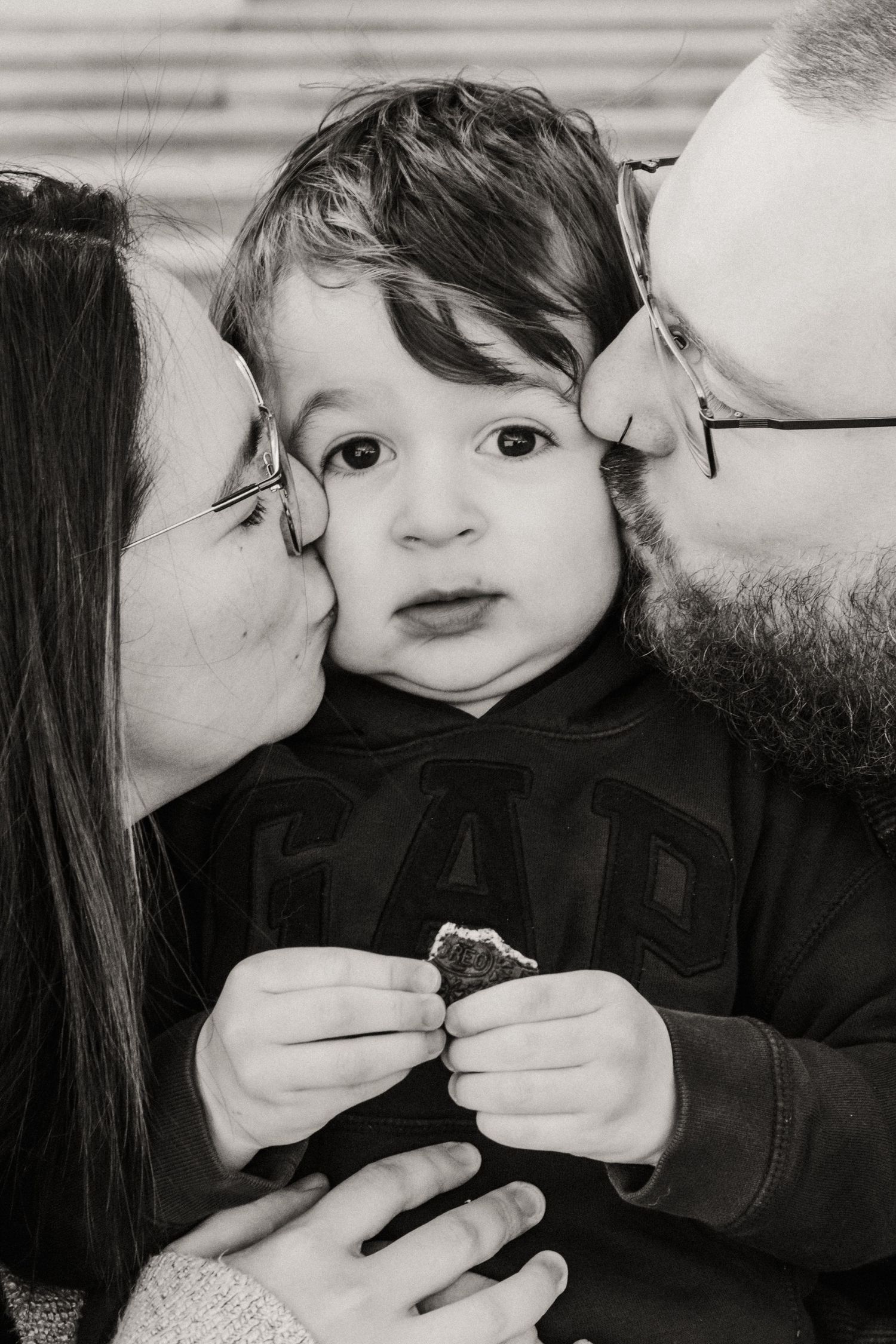 Black and white intimate family portrait showing parents giving kisses to a small child holding a treat.