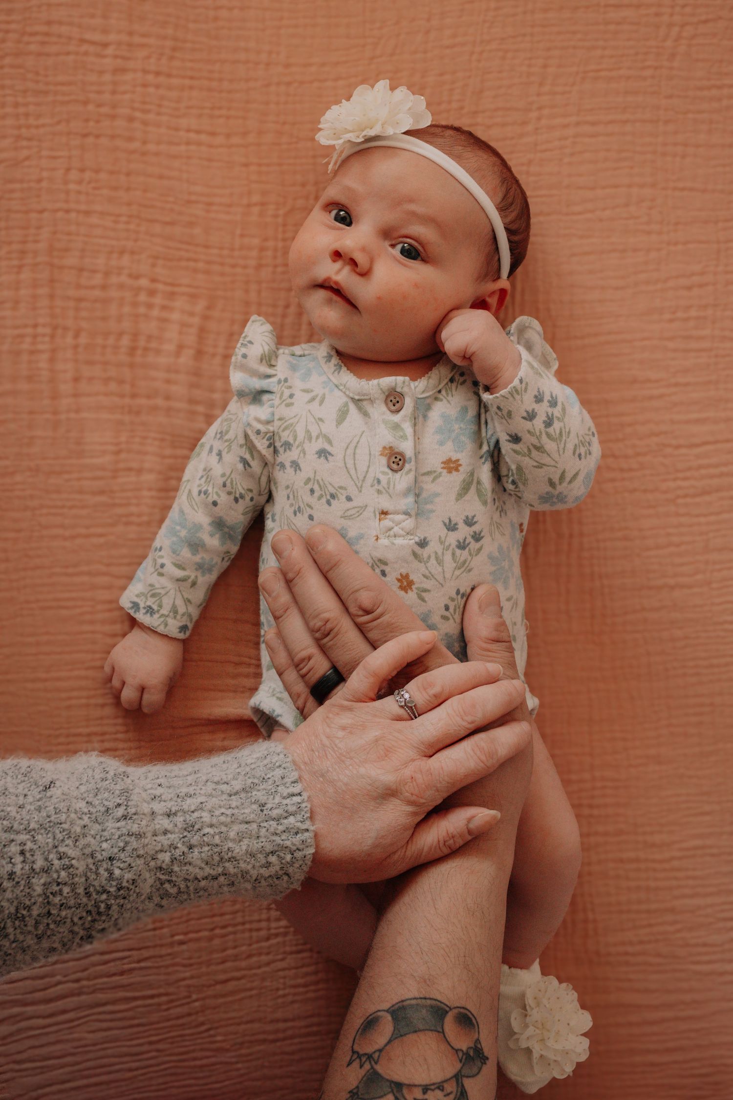 A baby wearing a floral onesie and white headband lies on a peach-colored backdrop while adult hands gently support.