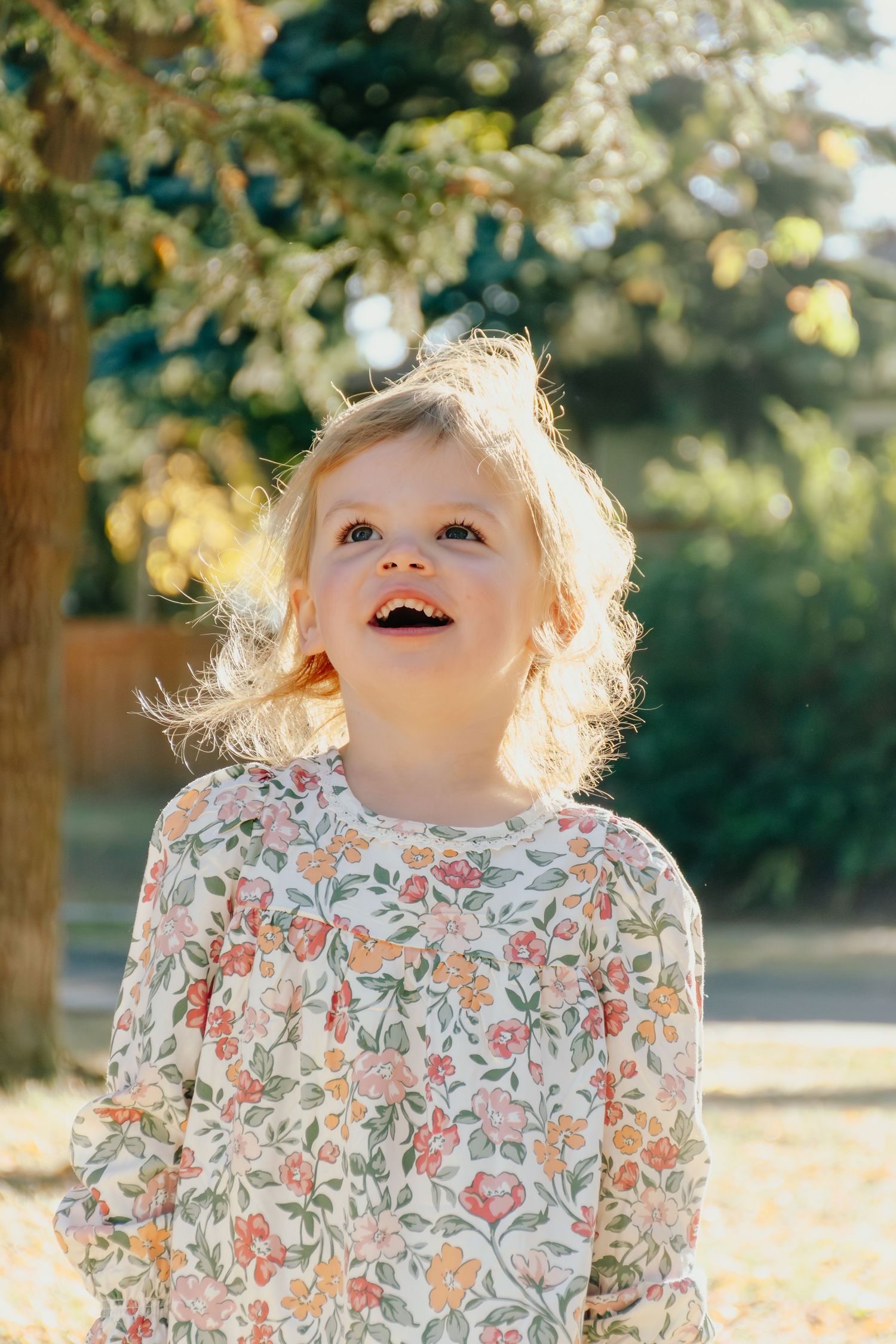 Young child in floral dress smiles joyfully while playing outdoors in golden sunlight.