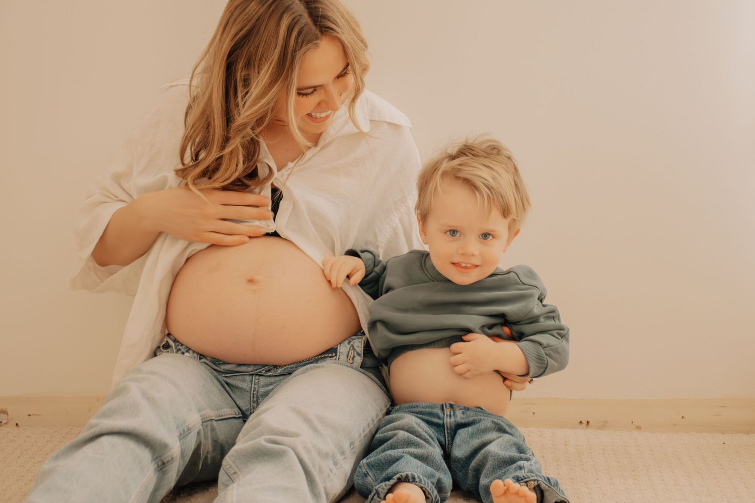 A pregnant woman and young child sit together on the floor, playfully showing their bare bellies.