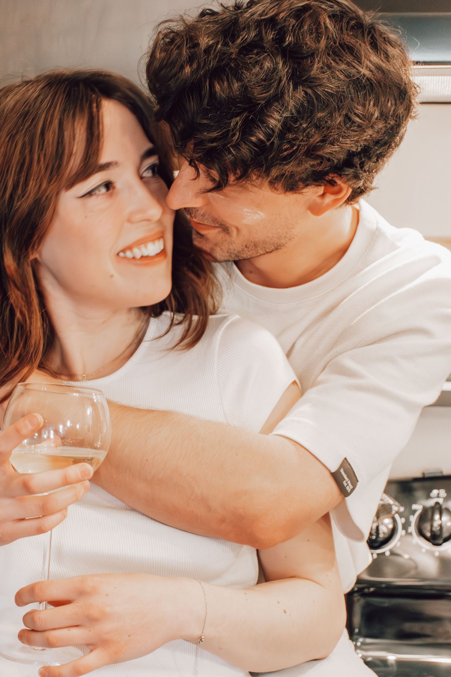 A romantic couple in white clothing share an intimate moment while holding a wine glass in their kitchen.
