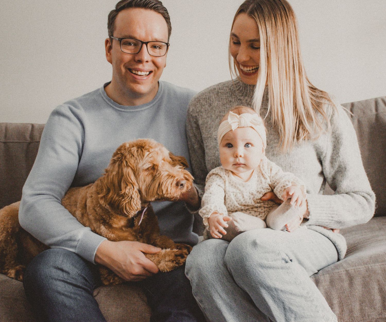 A family sits on a grey couch with their goldendoodle dog and baby in casual attire.