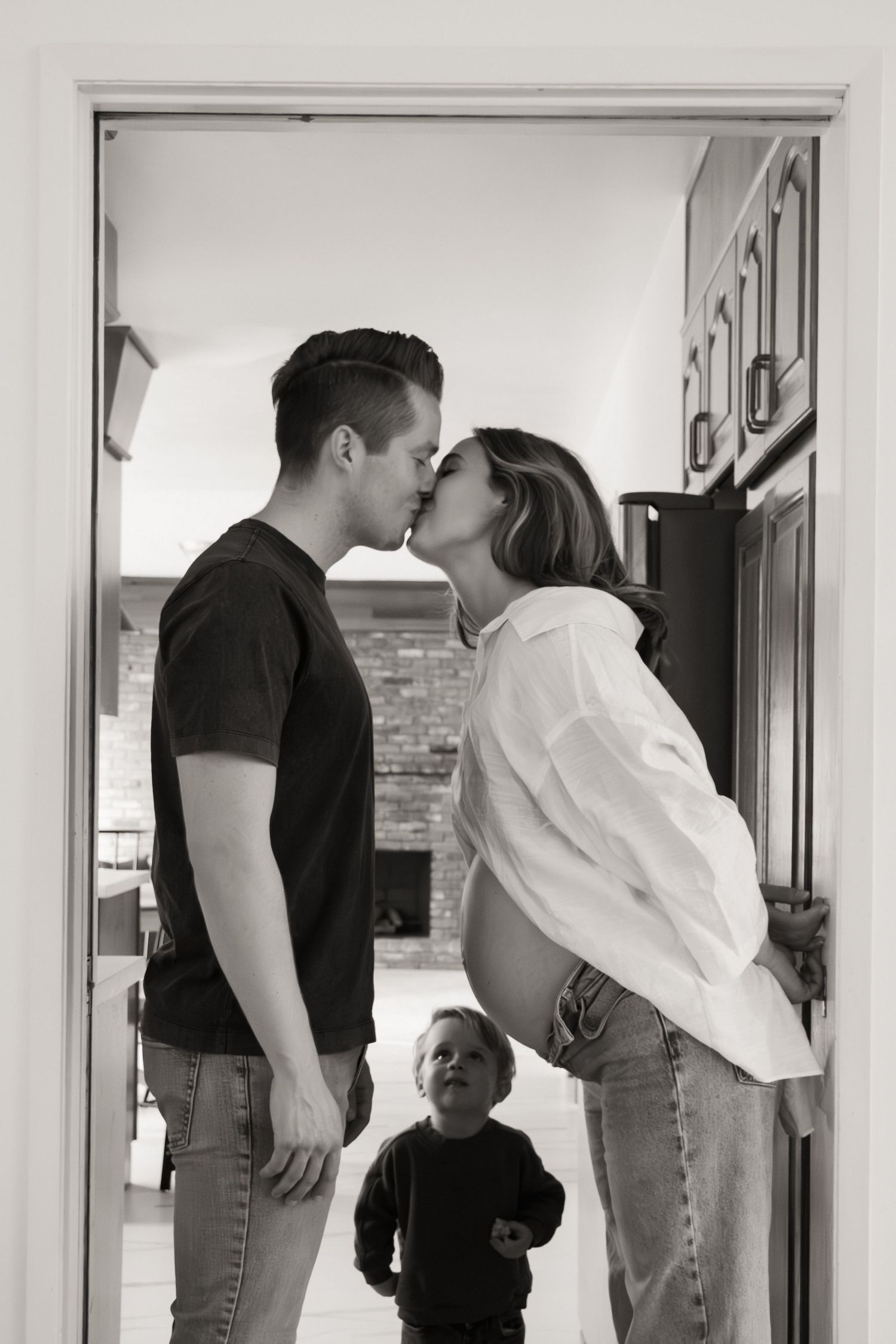 A couple shares an intimate kiss in a doorway while a young child in black watches from below.