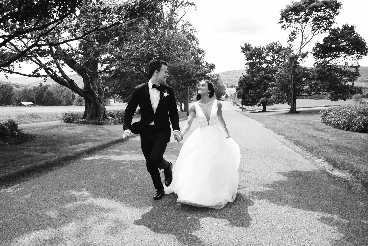 Bride and groom walking hand in hand along a tree-lined path in black and white.