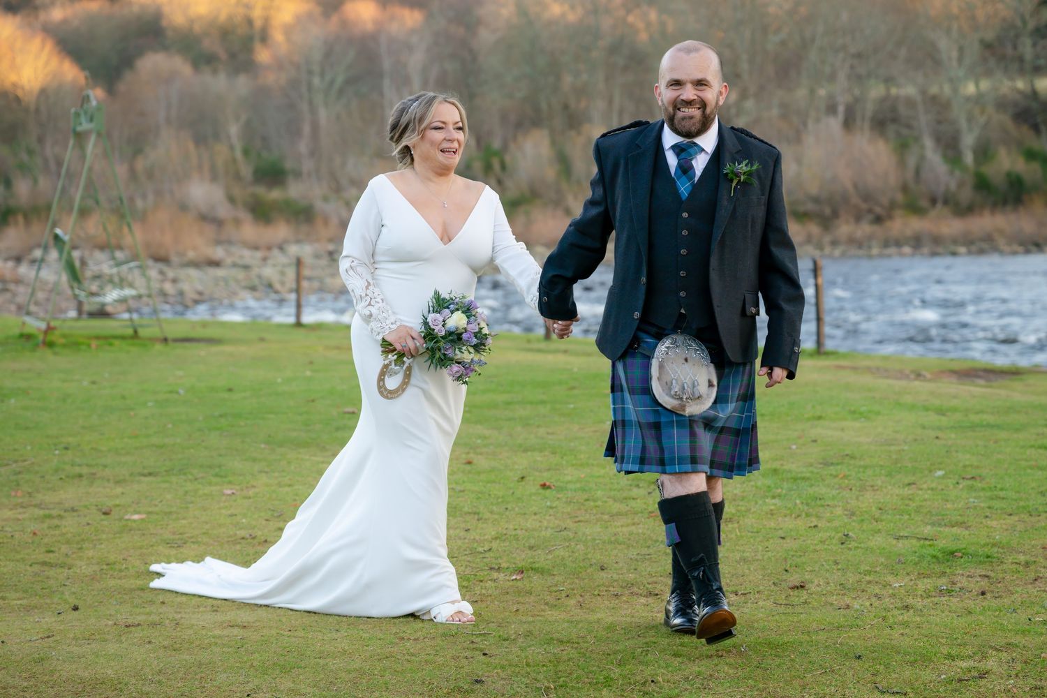 Wedding couple poses playfully in a grassy field near a lake at sunset during their autumn celebration.
