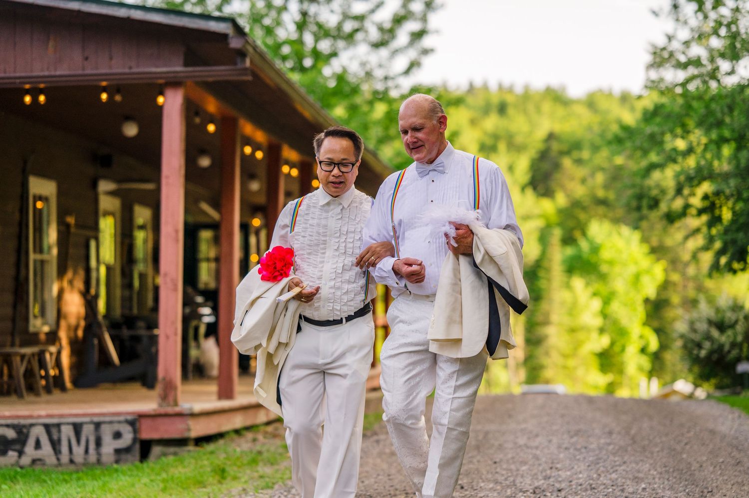 Two people in white attire walk together on a path near a camp building surrounded by lush greenery.