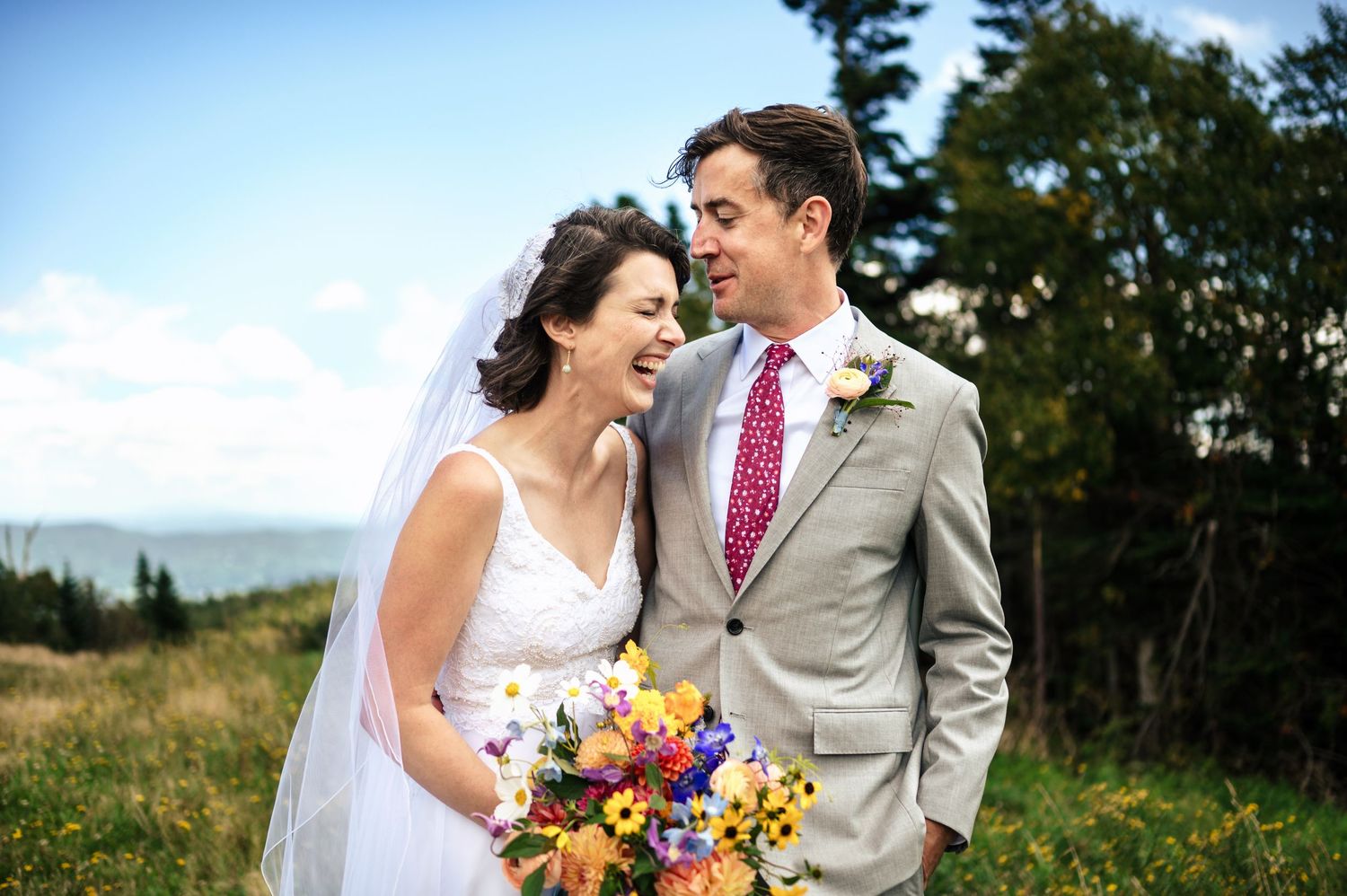 Smiling bride and groom share a joyful moment outdoors, she holds a colorful bouquet, he wears a gray suit with red tie.
