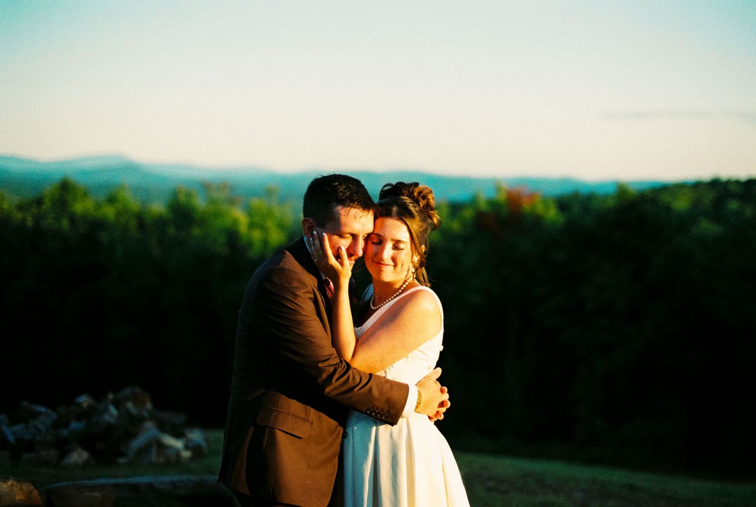 A couple embraces at golden hour with lush green mountains and clear blue sky in the background at their outdoor wedding.