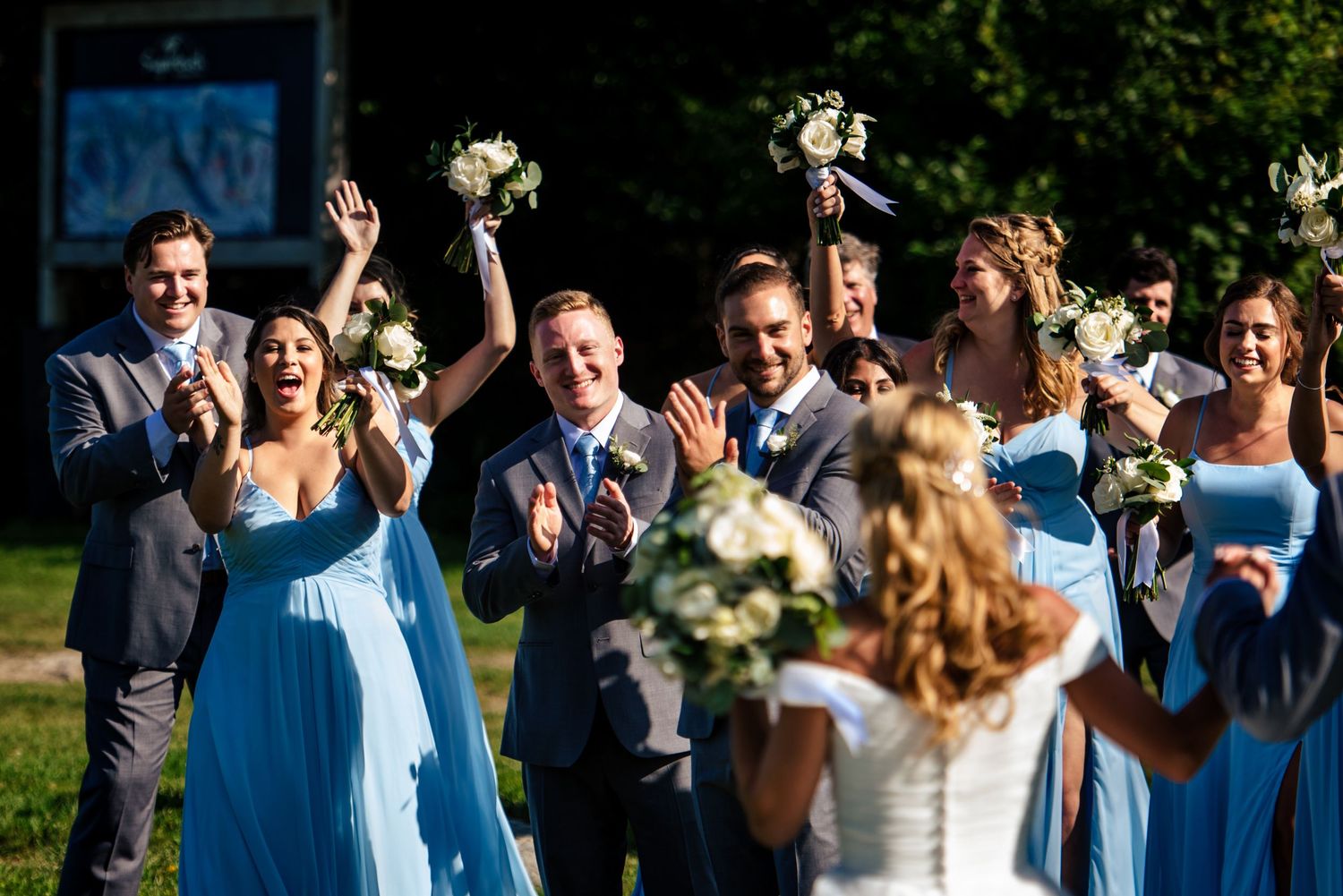 Wedding party in blue attire cheering for bride during outdoor bouquet toss celebration on sunny day.