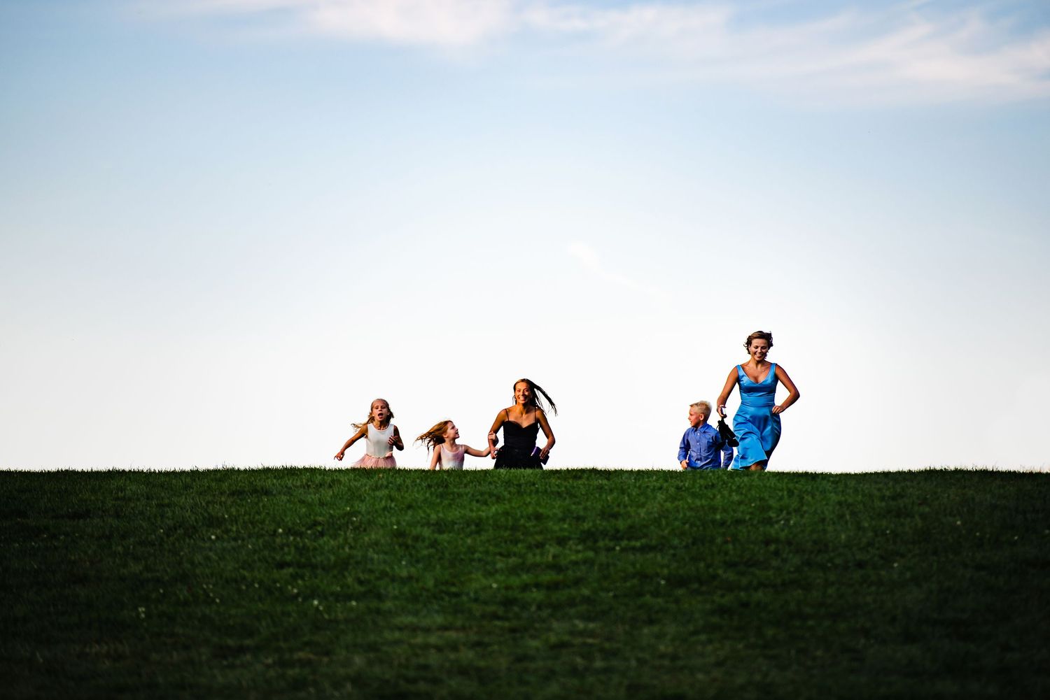 Five children running and playing on a lush green hill under a clear blue sky on a sunny day.