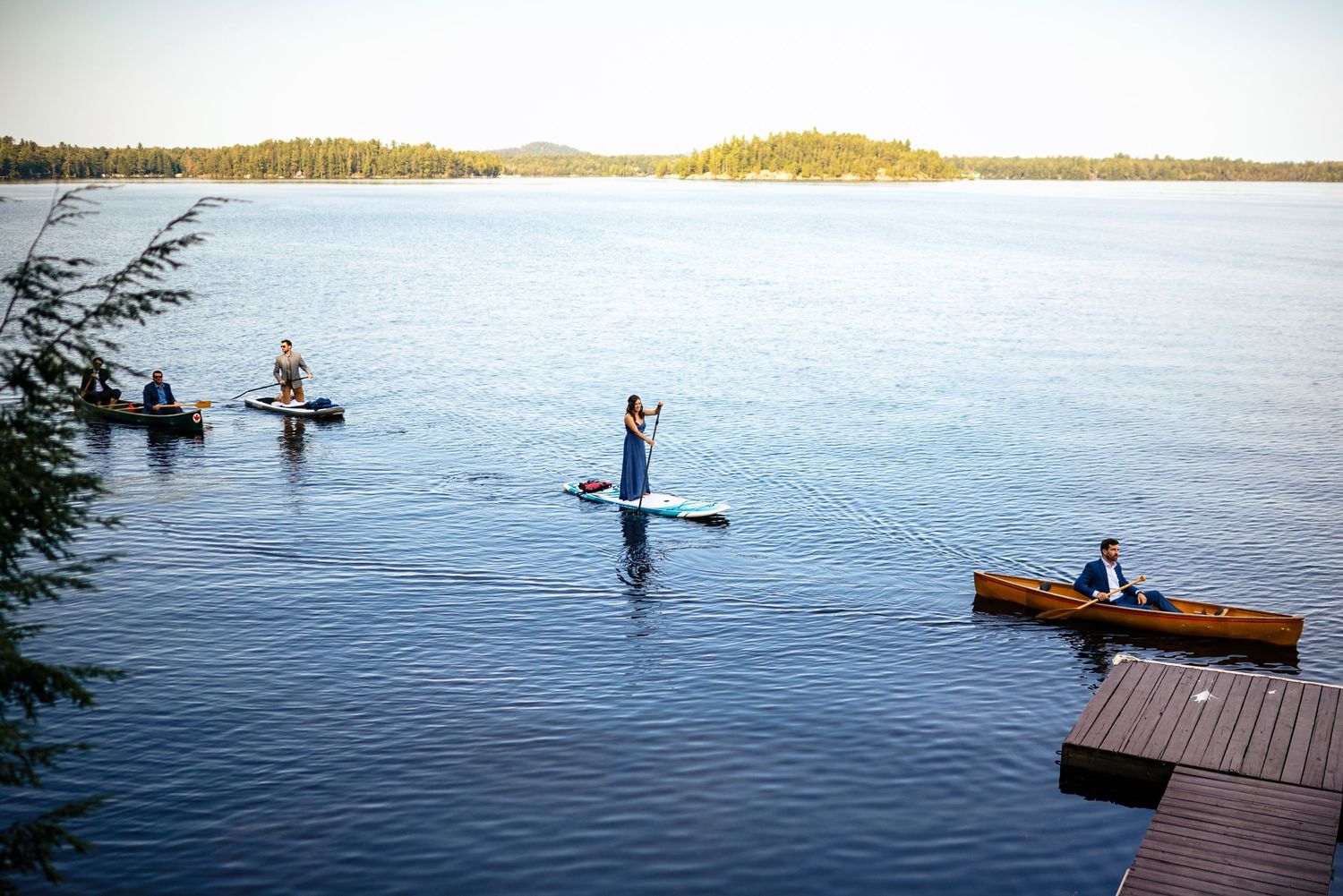 People paddleboarding and kayaking on a calm lake near a wooden dock with forested shoreline in the background.