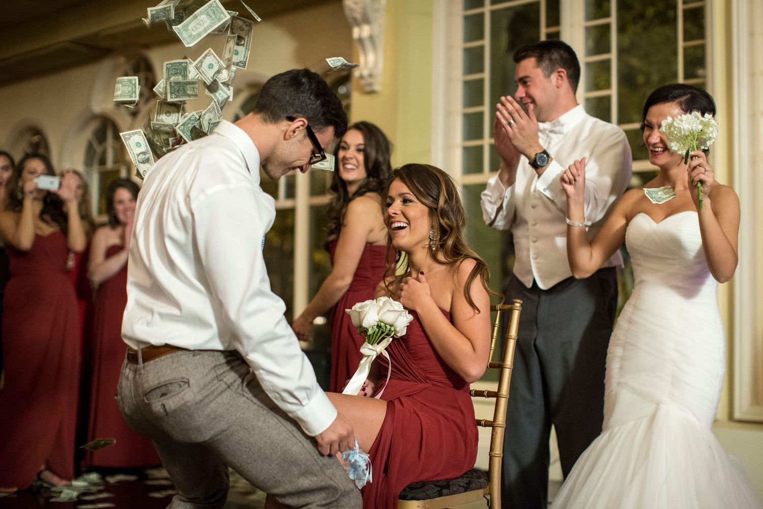 Groom removes garter from bride as money flies through the air at a lively wedding reception with bridesmaids.