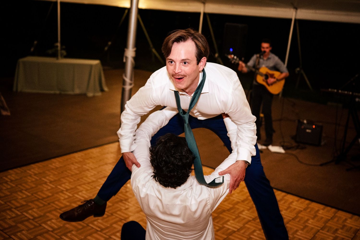 Two men dancing energetically at a wedding reception under a tent, with a guitarist performing in the background.