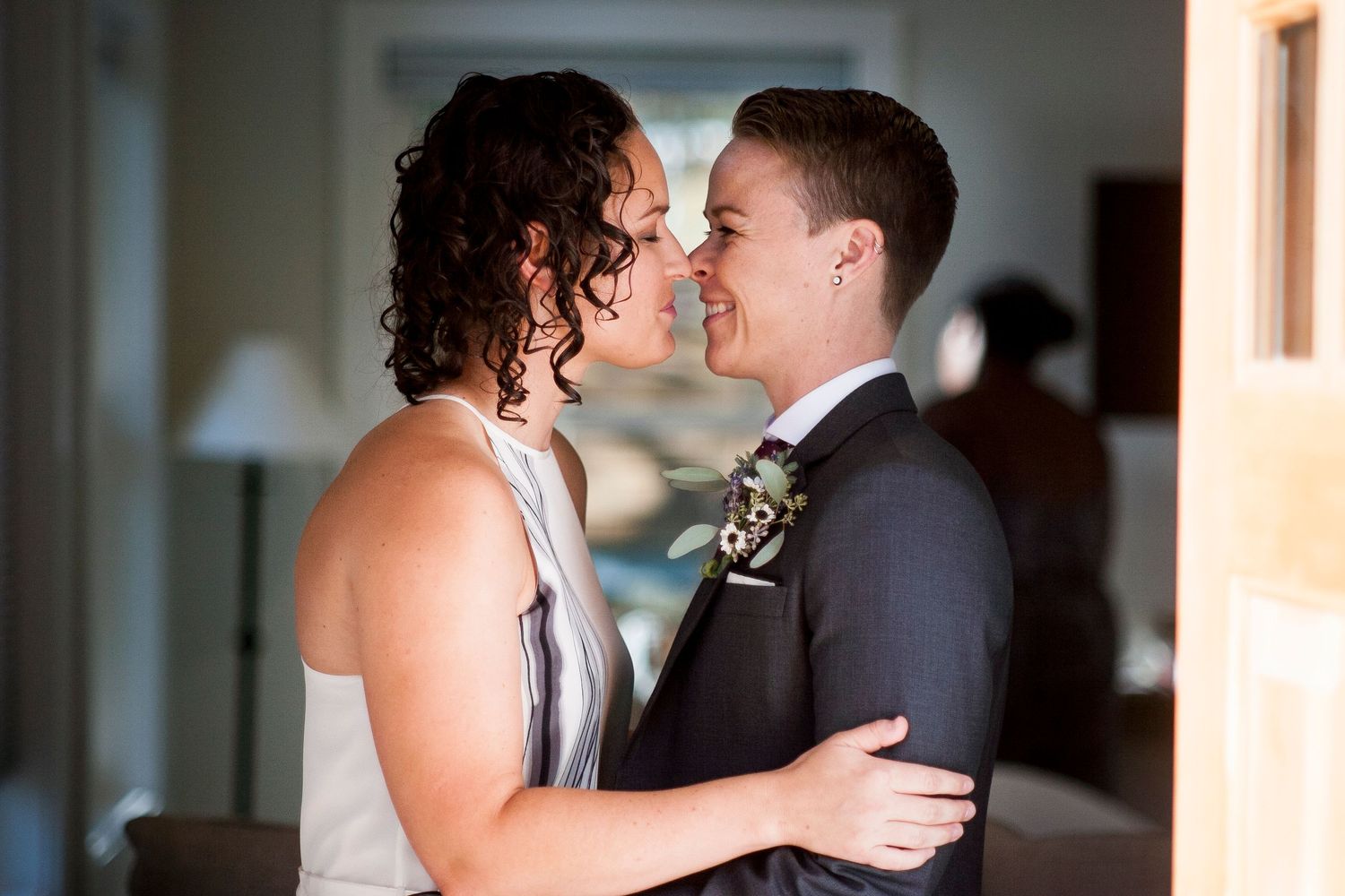 Two brides sharing a tender moment, one in a white dress and one in a dark suit with boutonniere, on their wedding day.