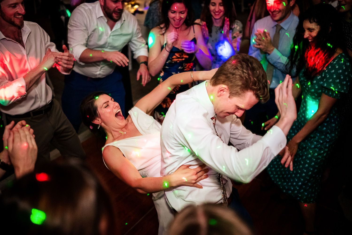Bride and groom dancing energetically at wedding reception party with colorful lights and guests celebrating in background.