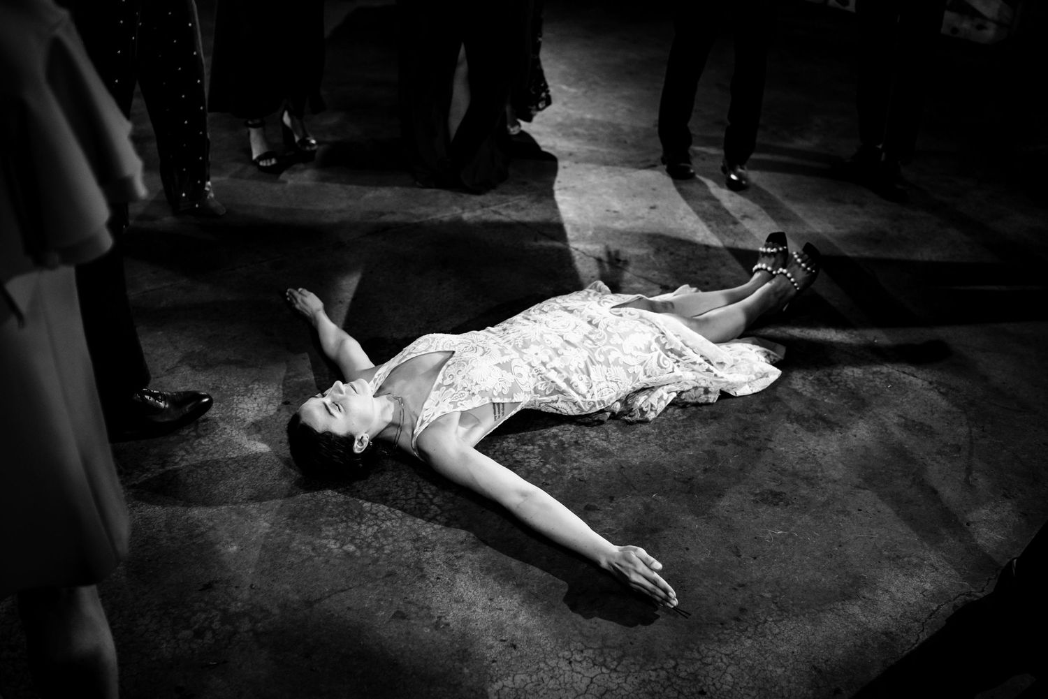 Black and white photo of a woman in a white dress lying on a dance floor surrounded by standing guests.